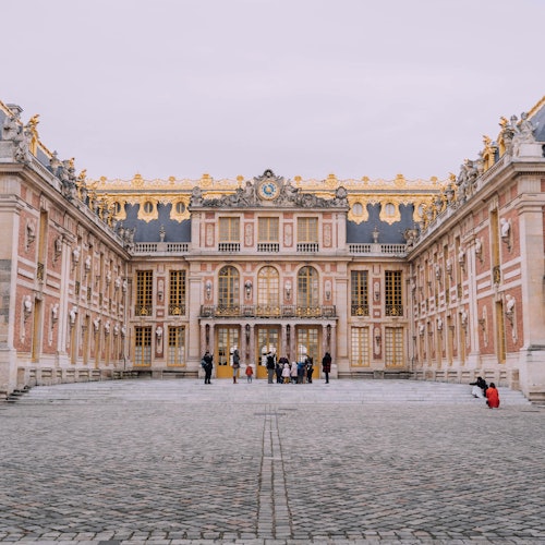 People standing in a cobblestone courtyard in front of an ornate, symmetrical palace with golden details and a clock.