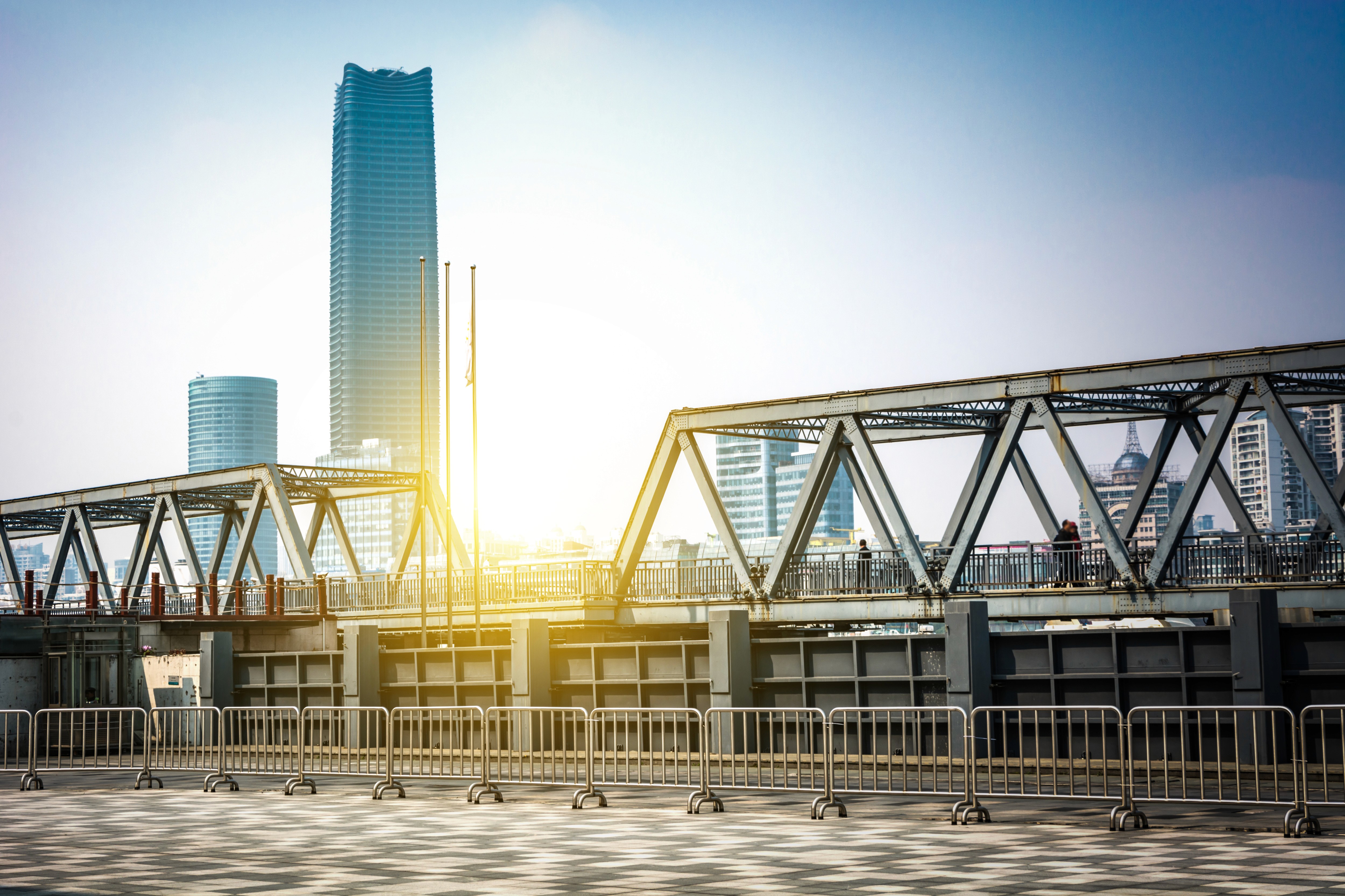 A sunlit cityscape with a modern steel bridge in the foreground. Tall skyscrapers rise in the background under a clear blue sky, creating an urban feel.