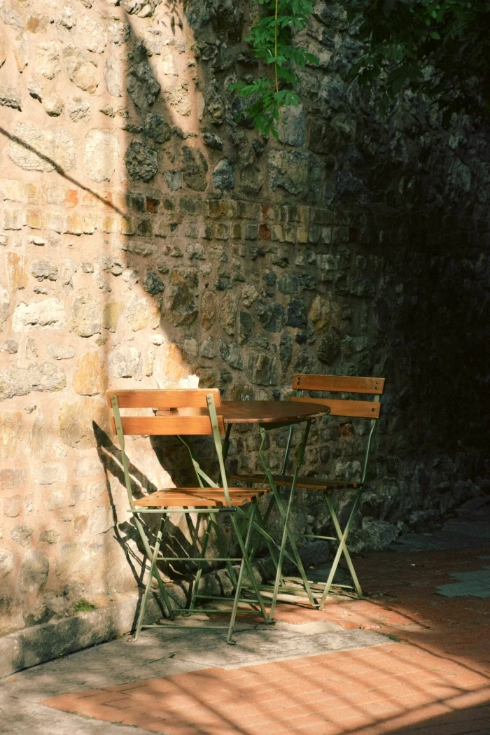 Two wooden chairs at a table beside a sun-warmed stone wall.