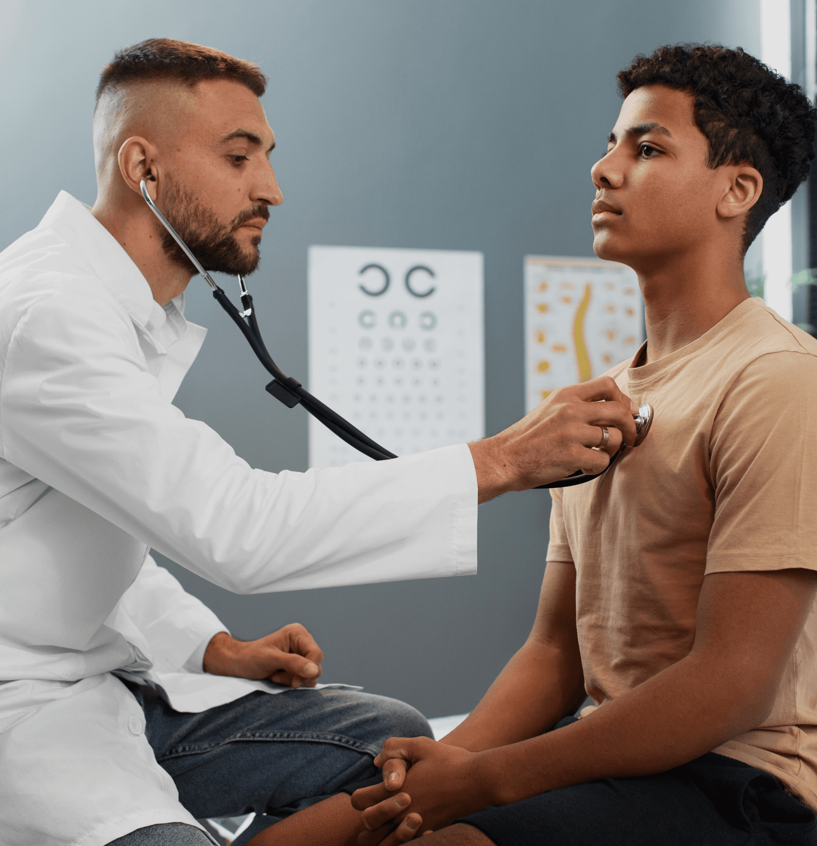 A doctor in a white coat uses a stethoscope to listen to a teenage boy's chest in a medical office. Eye charts are visible in the background.