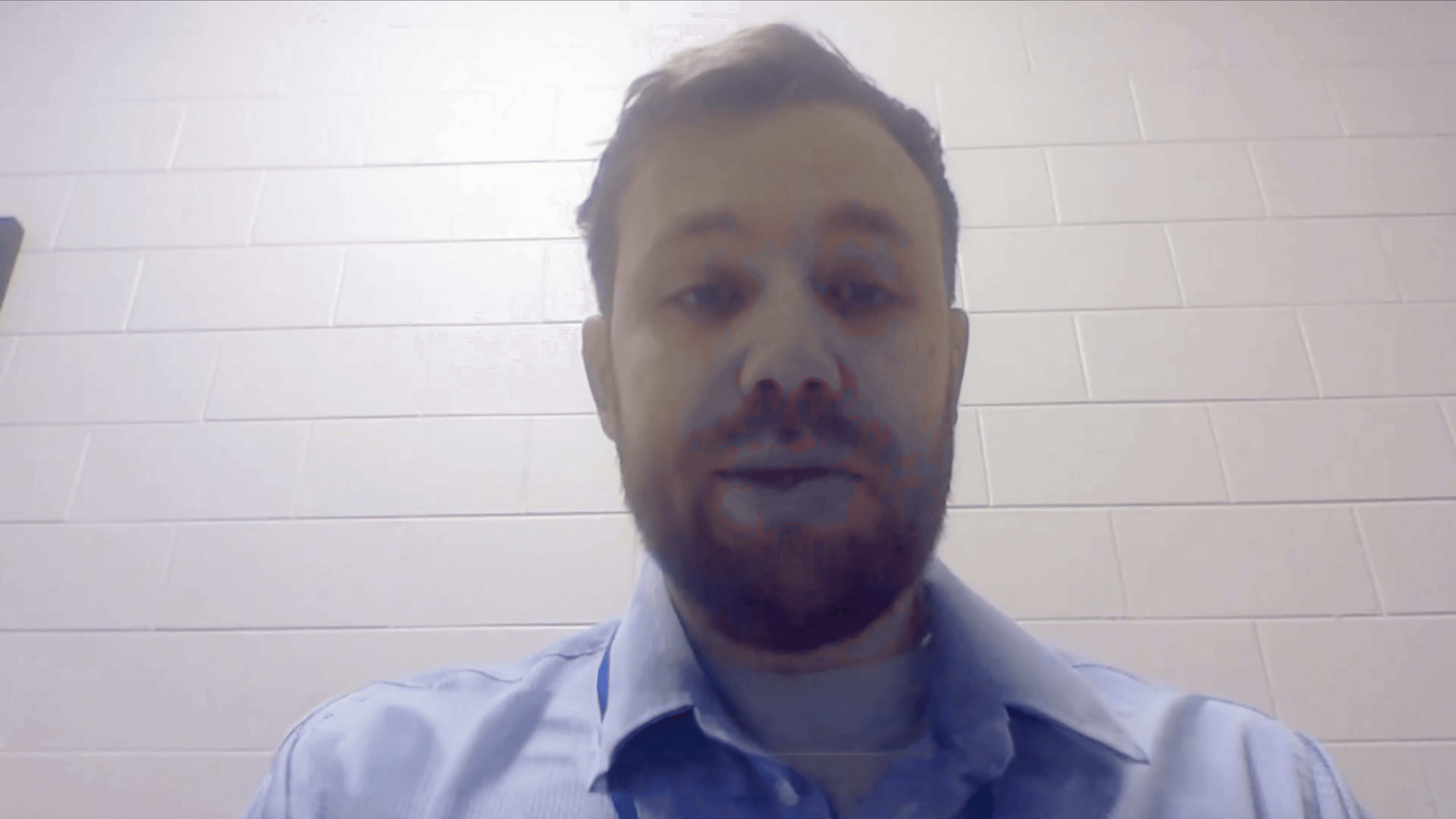 A man with short hair and a trimmed beard appears on camera, facing forward as if in a video call or recording. He is wearing a light blue collared shirt. The background shows a plain, light-colored brick wall, giving the setting a simple indoor environment such as an office or classroom.