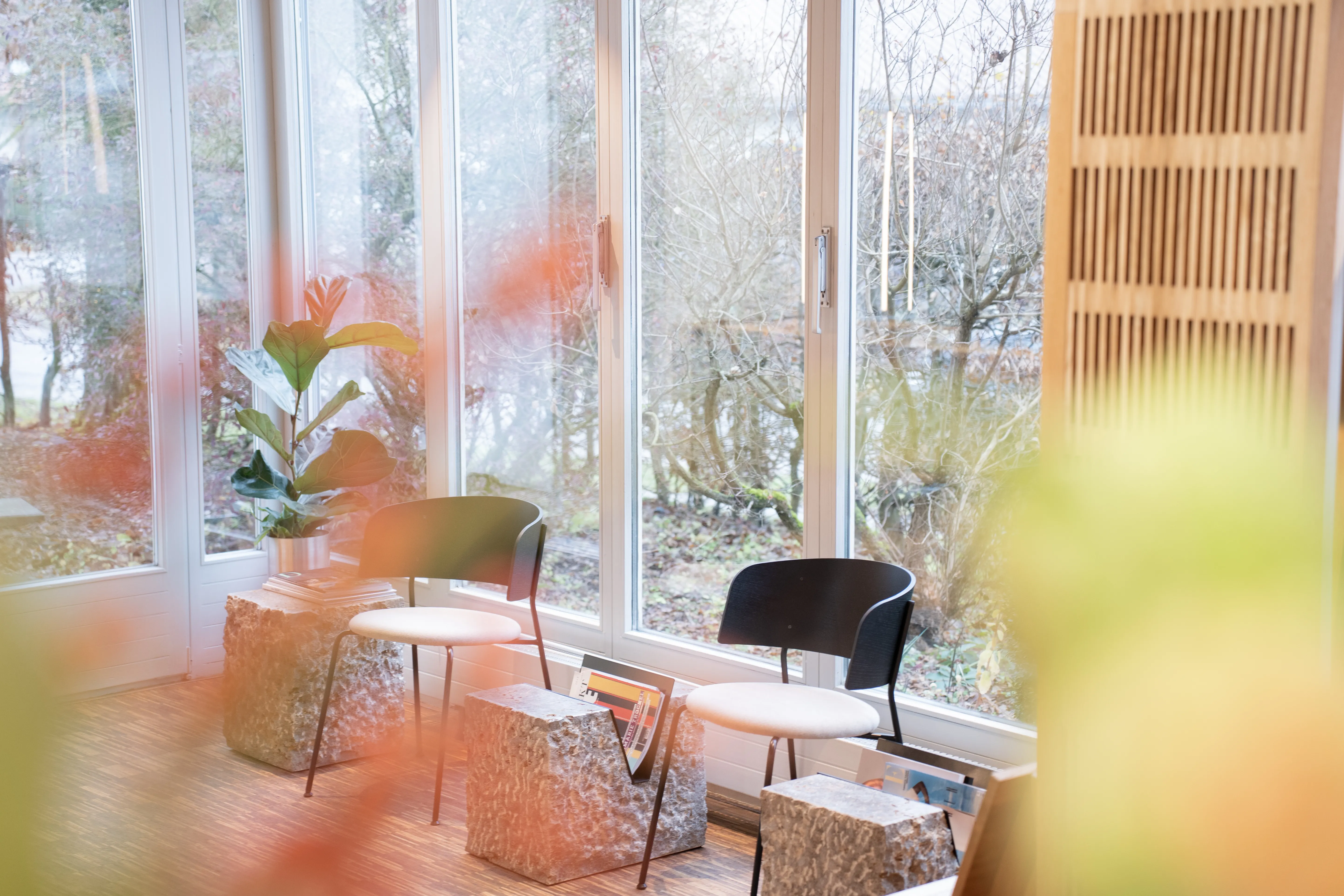 Window lounge at EMPL FRISEURE with two chairs in black and cream, stone-look side cubes with magazines and a fig plant in front of large windows facing the garden.