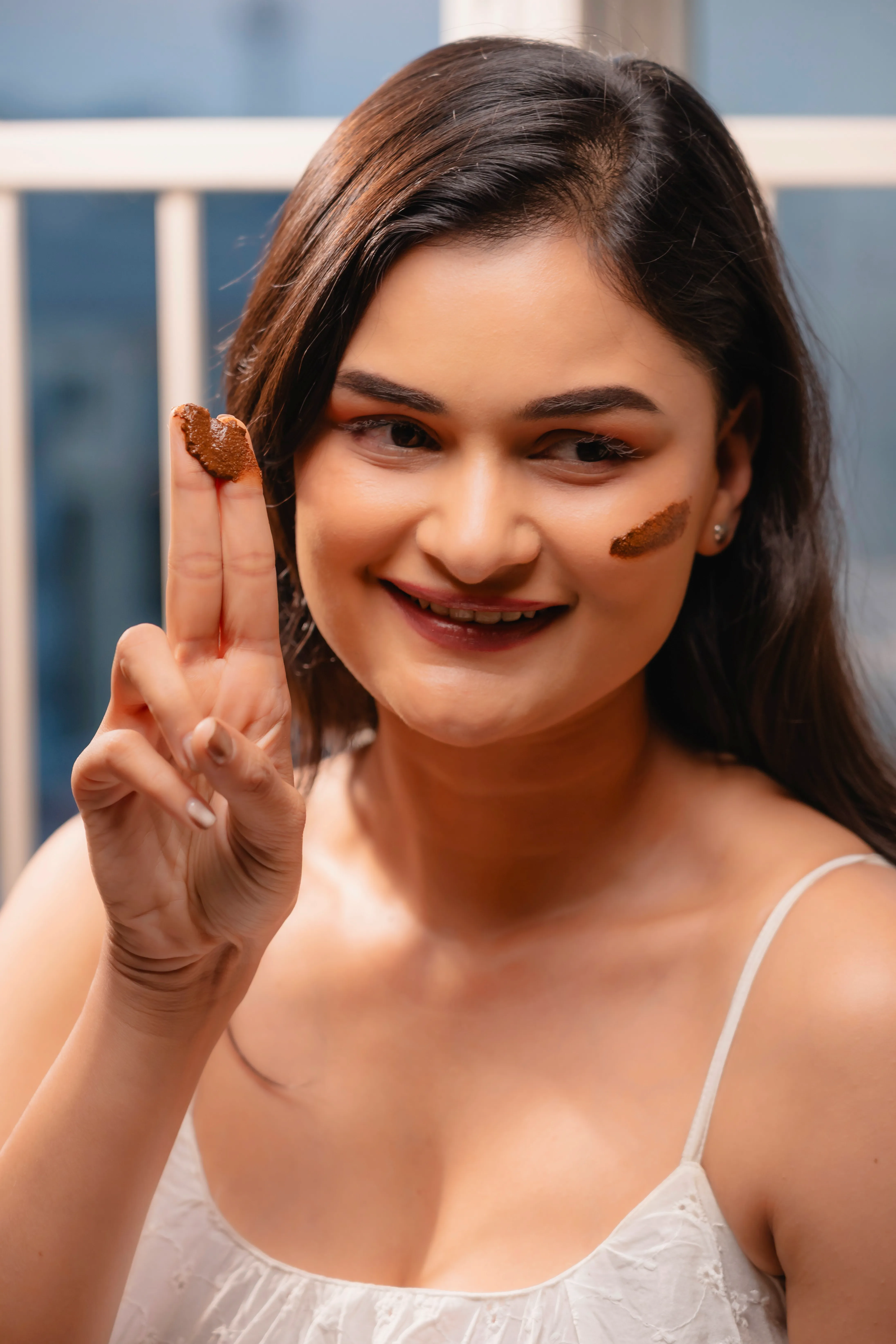 A person smiling, raising two fingers in a peace sign, wearing a light dress, with a soft background.