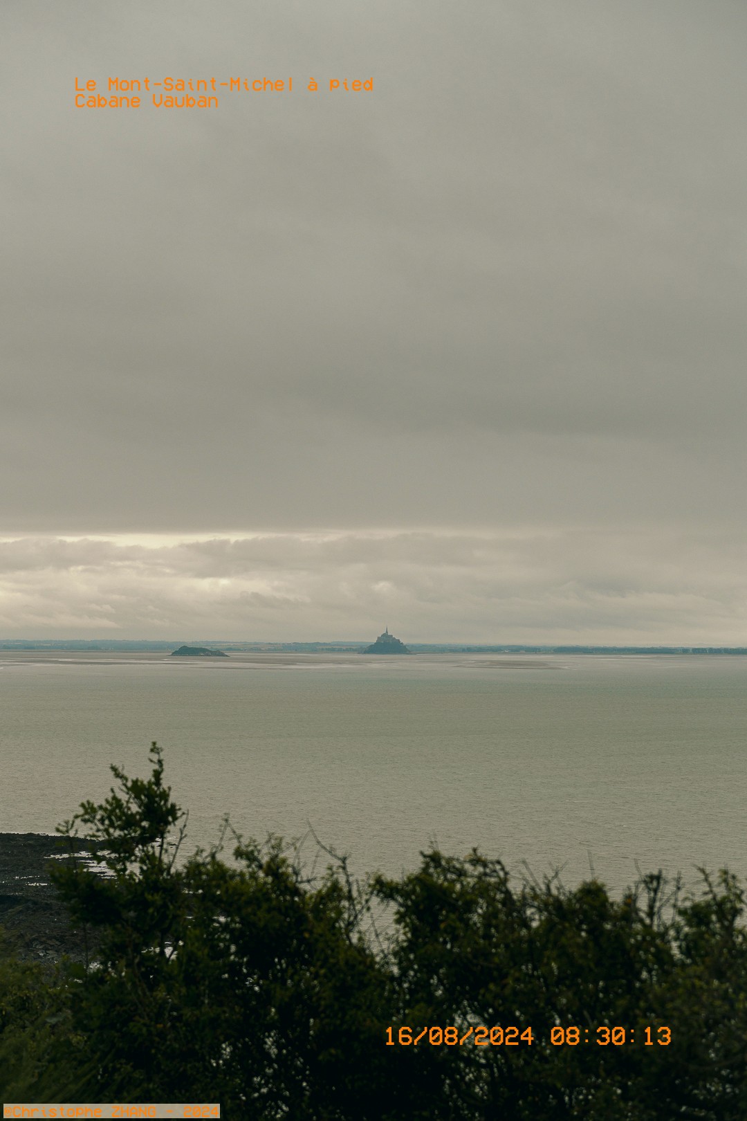 Scène de randonnée côtière, une marcheuse observant la baie du Mont-Saint-Michel sous un ciel couvert.