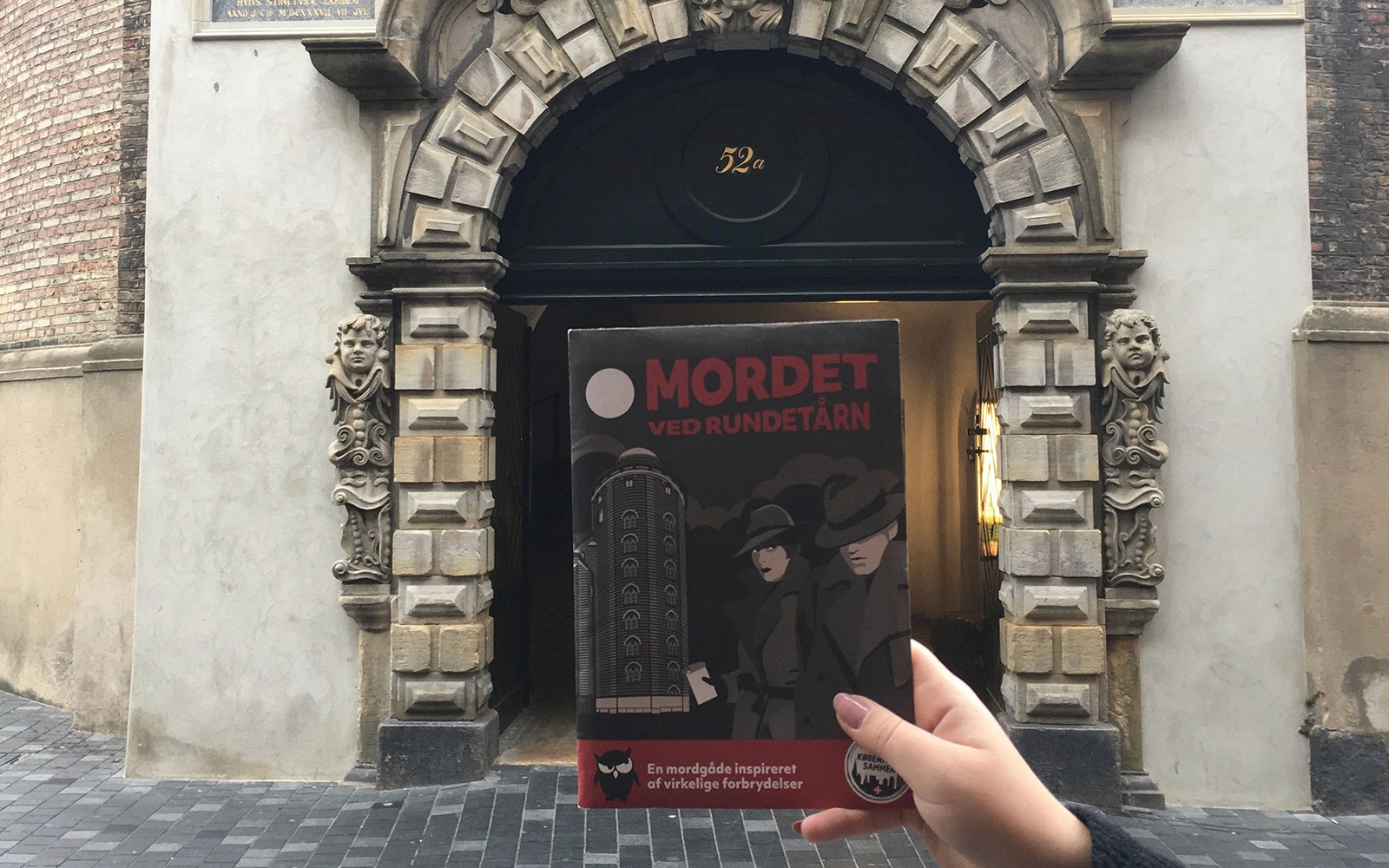 Gyilkossági Rejtély Élmény prospektus tartva a Kerek Torony bejárata előtt, Koppenhága.