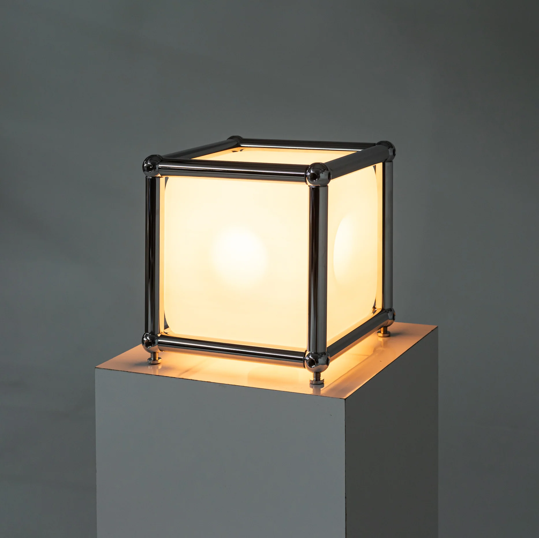 Rarify Cube One table lamp lit, translucent cube panels held in a chrome USM Haller frame on a brass plate base