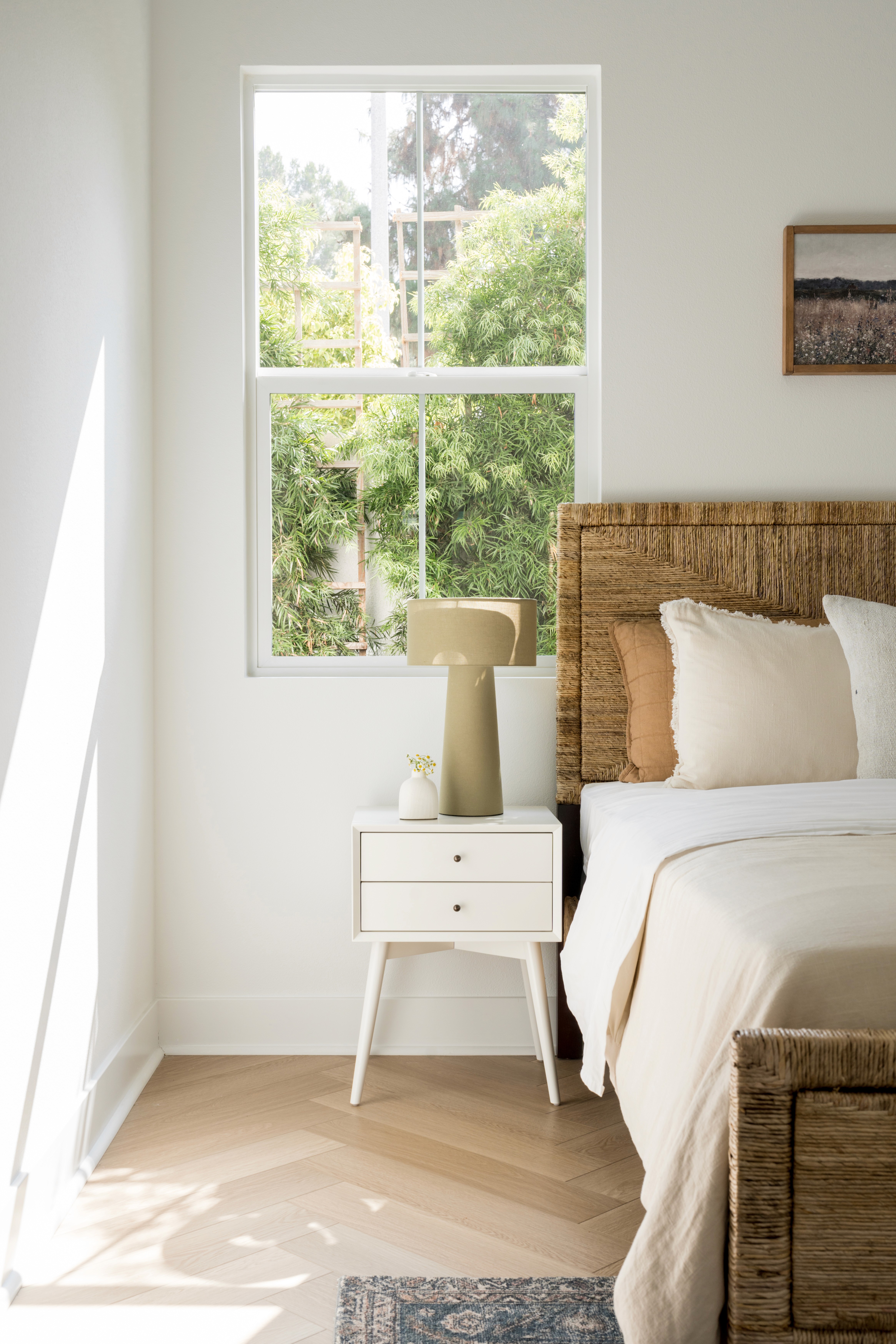 Guest bedroom with natural light, chevron flooring, woven bed frame, white two-drawer side table, and large table lamp by Jubilee Interiors in Arcadia, California