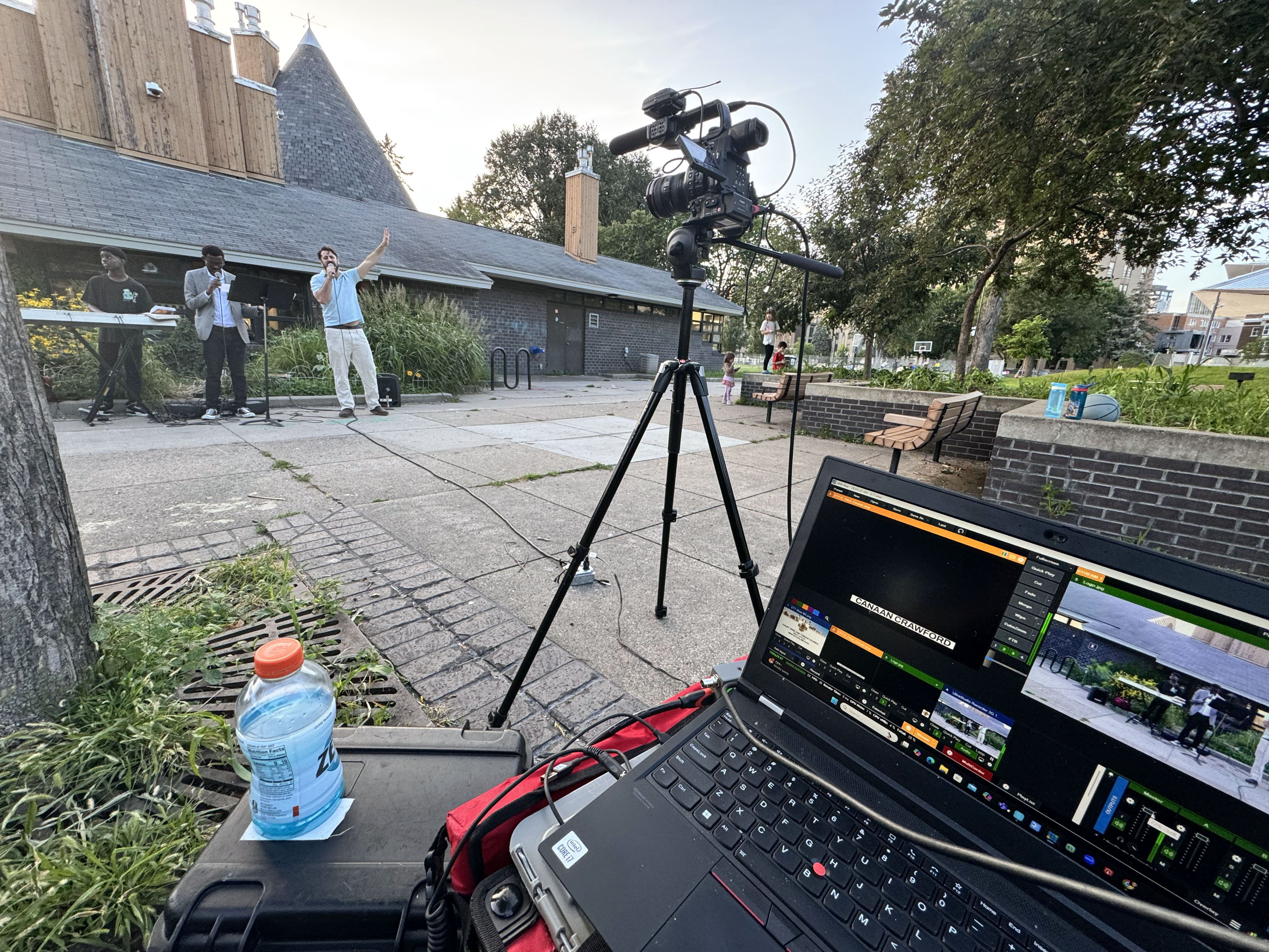 A laptop running livestream software during an outdoor musical performance in a paved public park setting.