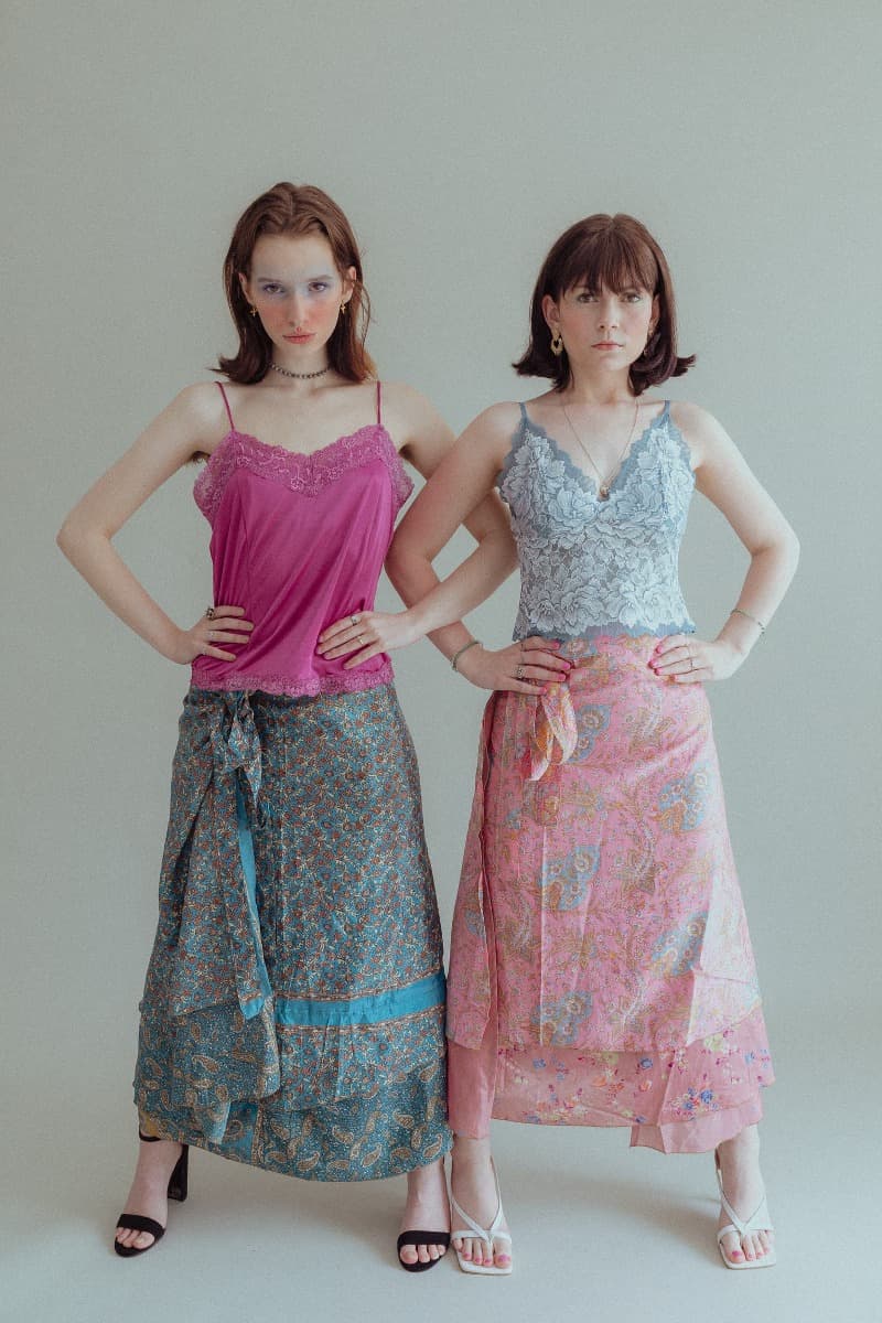 Two models standing side by side, one wearing a pink top and floral skirt, the other in a gray lace top and pink skirt, captured in an ethereal photoshoot at Revelator Studio, a natural light photography studio in Shreveport.