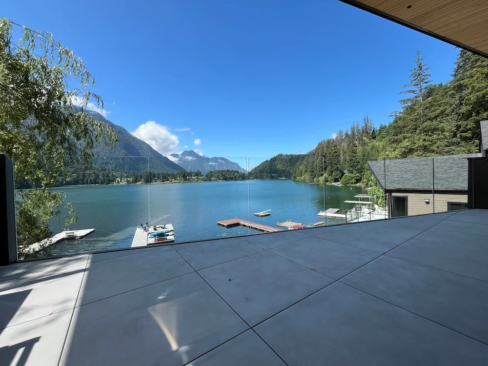 Expansive lakefront view framed by frameless glass railing on an elevated patio, offering clear mountain and water vistas in British Columbia.