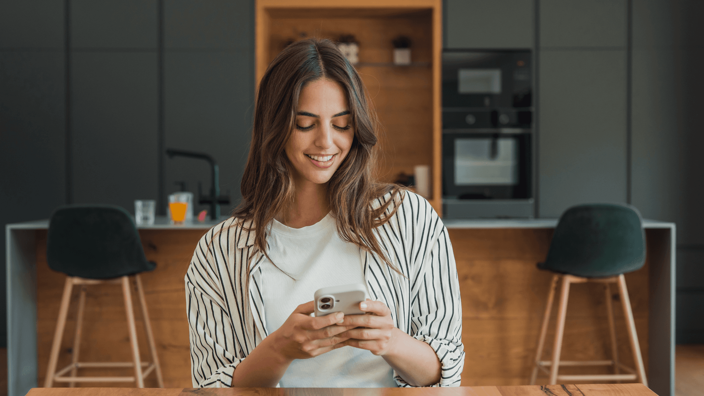 Smiling woman in a striped shirt checking her phone while sitting at a kitchen counter, representing a client staying connected during her recovery