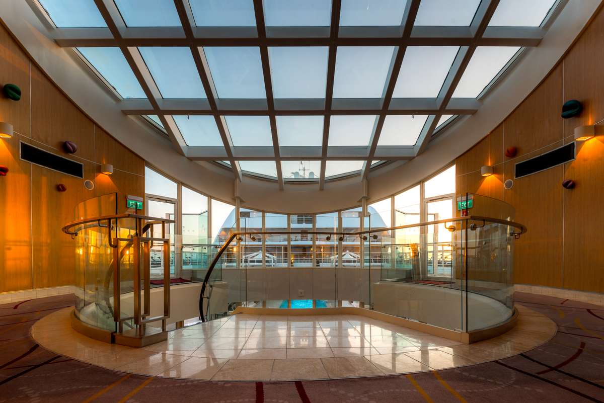 A modern spa interior features a glass ceiling, warm wood paneling, and two curved glass partitions over a pool area.