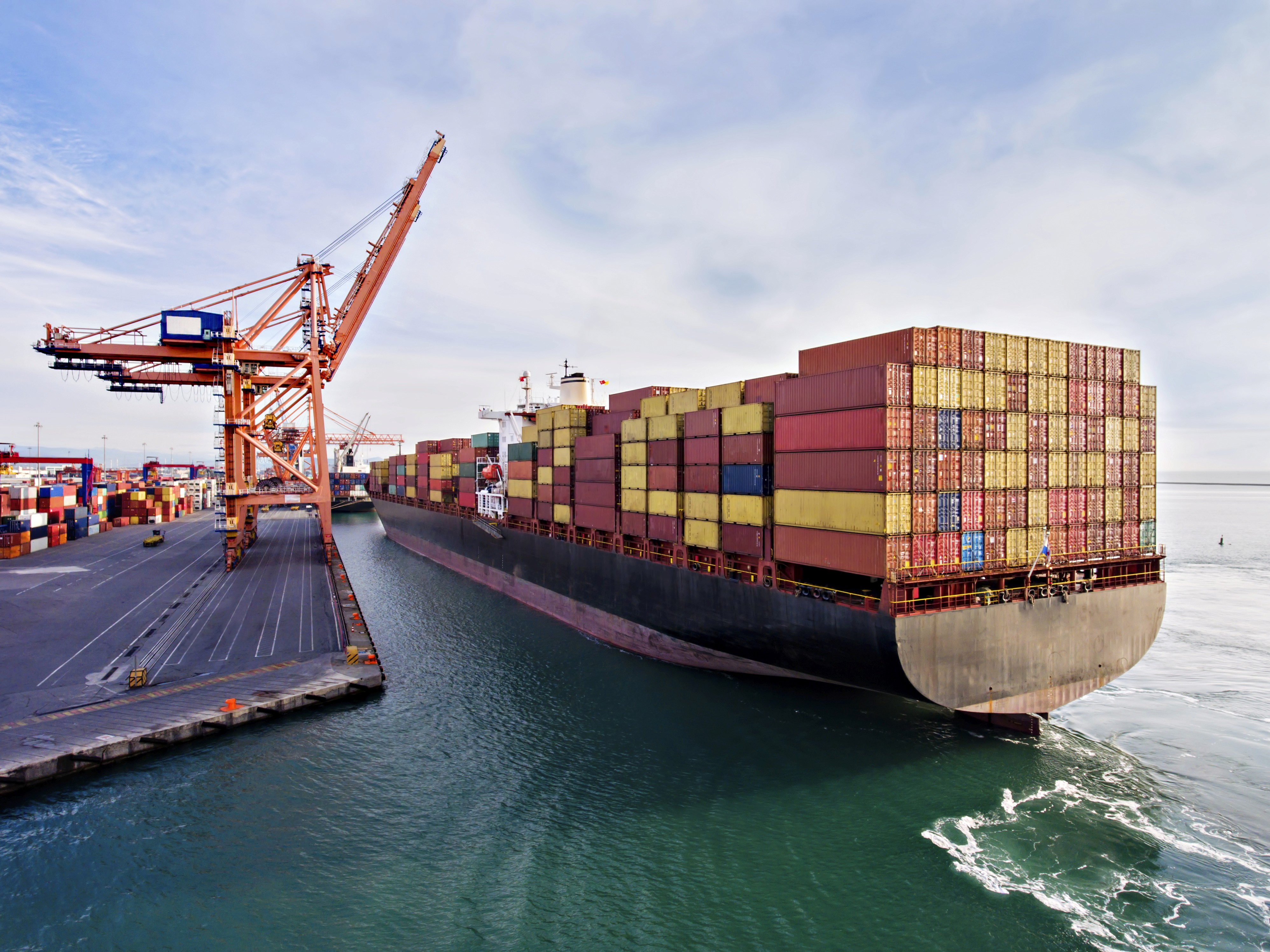 A large cargo ship with stacks of colorful shipping containers is docked at an industrial port, adjacent to a towering orange crane under a clear blue sky.