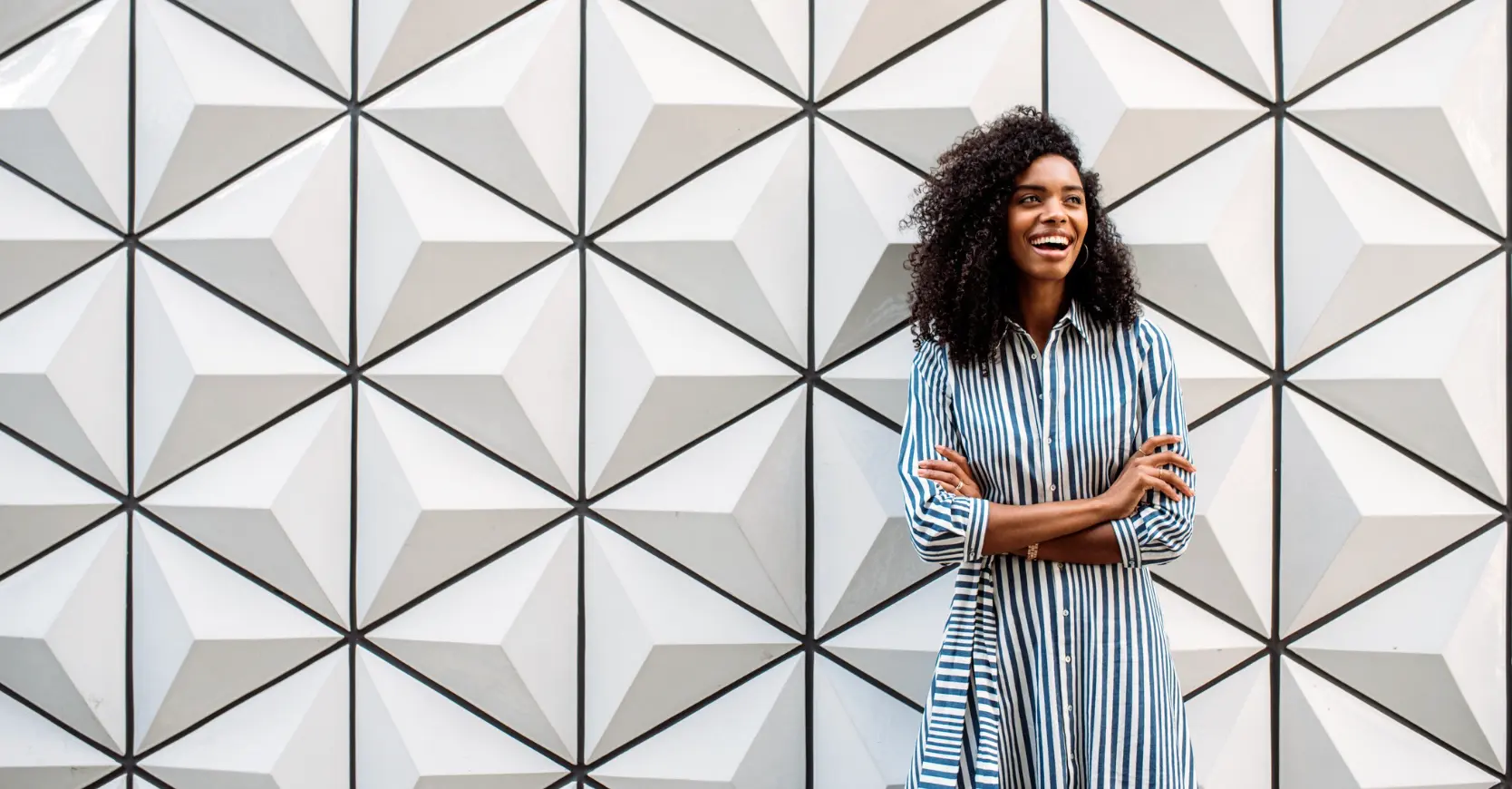 Confident woman laughing with arms crossed against a geometric triangular wall pattern, representing personal strategy and an aspirational future vision