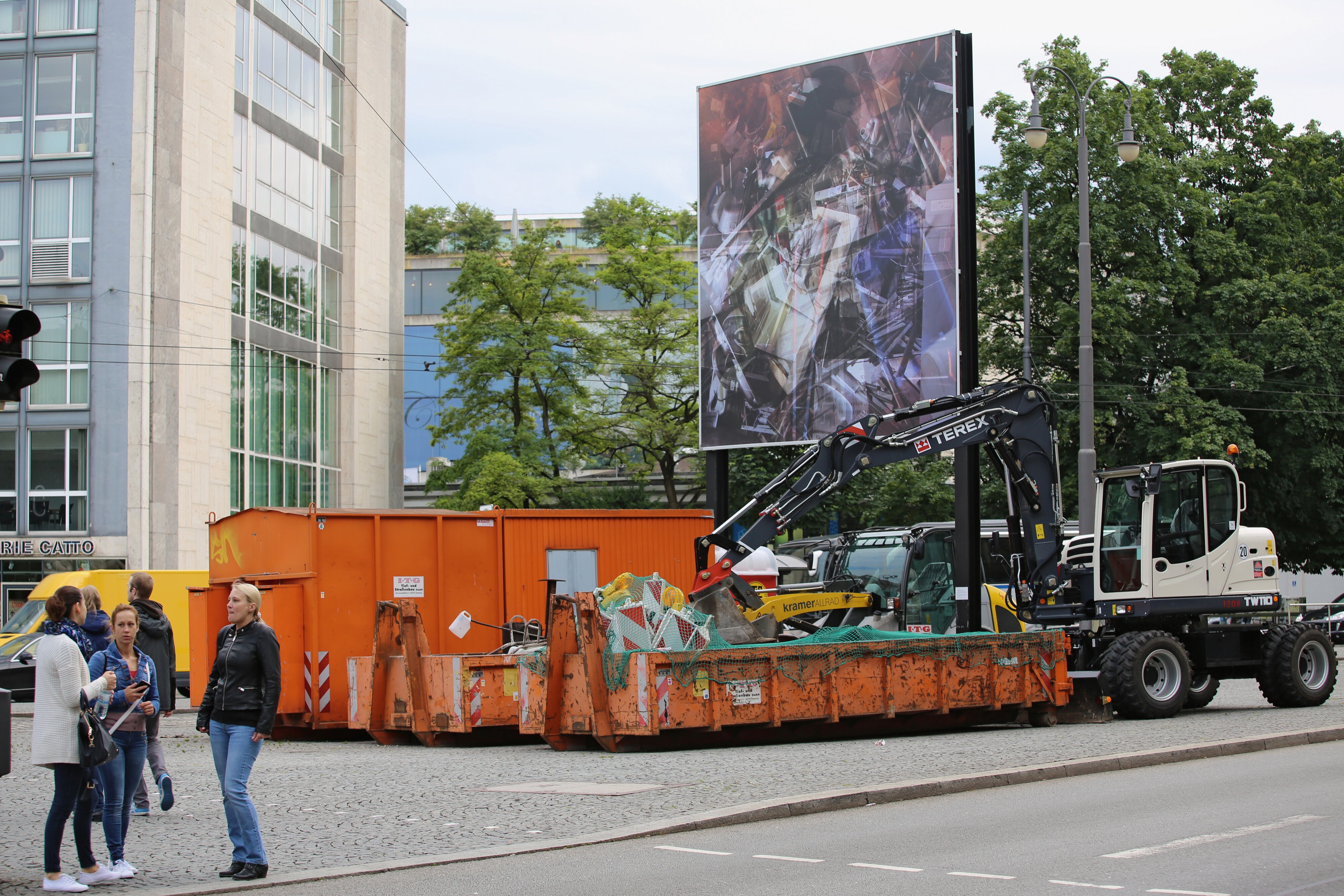 Bildwand mit der Collage am Lenbachplatz, davor orange Container, ein Bagger und Passanten