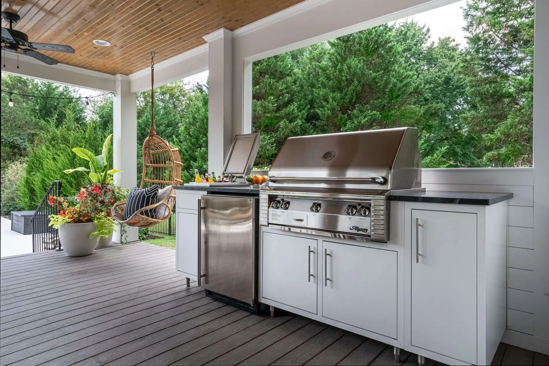 A stylish outdoor kitchen with white cabinetry and a stainless steel grill, complemented by a mini-fridge, under a wood-paneled ceiling. A hanging rattan chair and lush greenery add comfort and elegance to the patio.