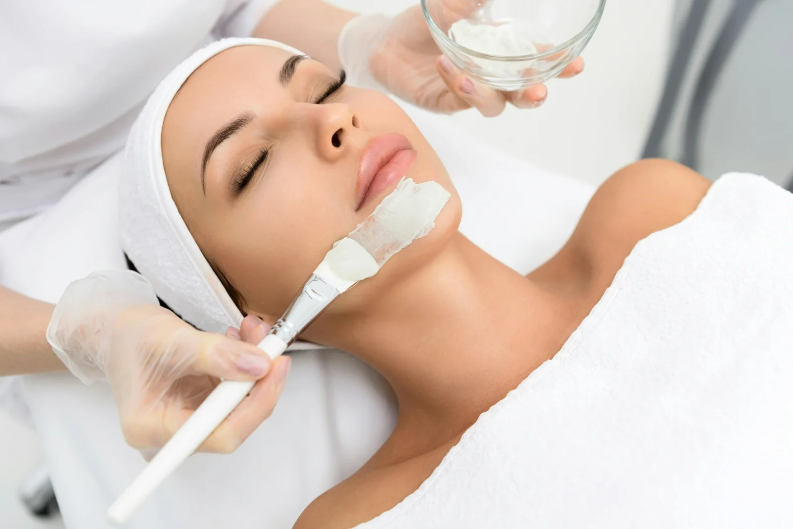 Esthetician applies white facial mask to relaxed client's face during professional skincare treatment.