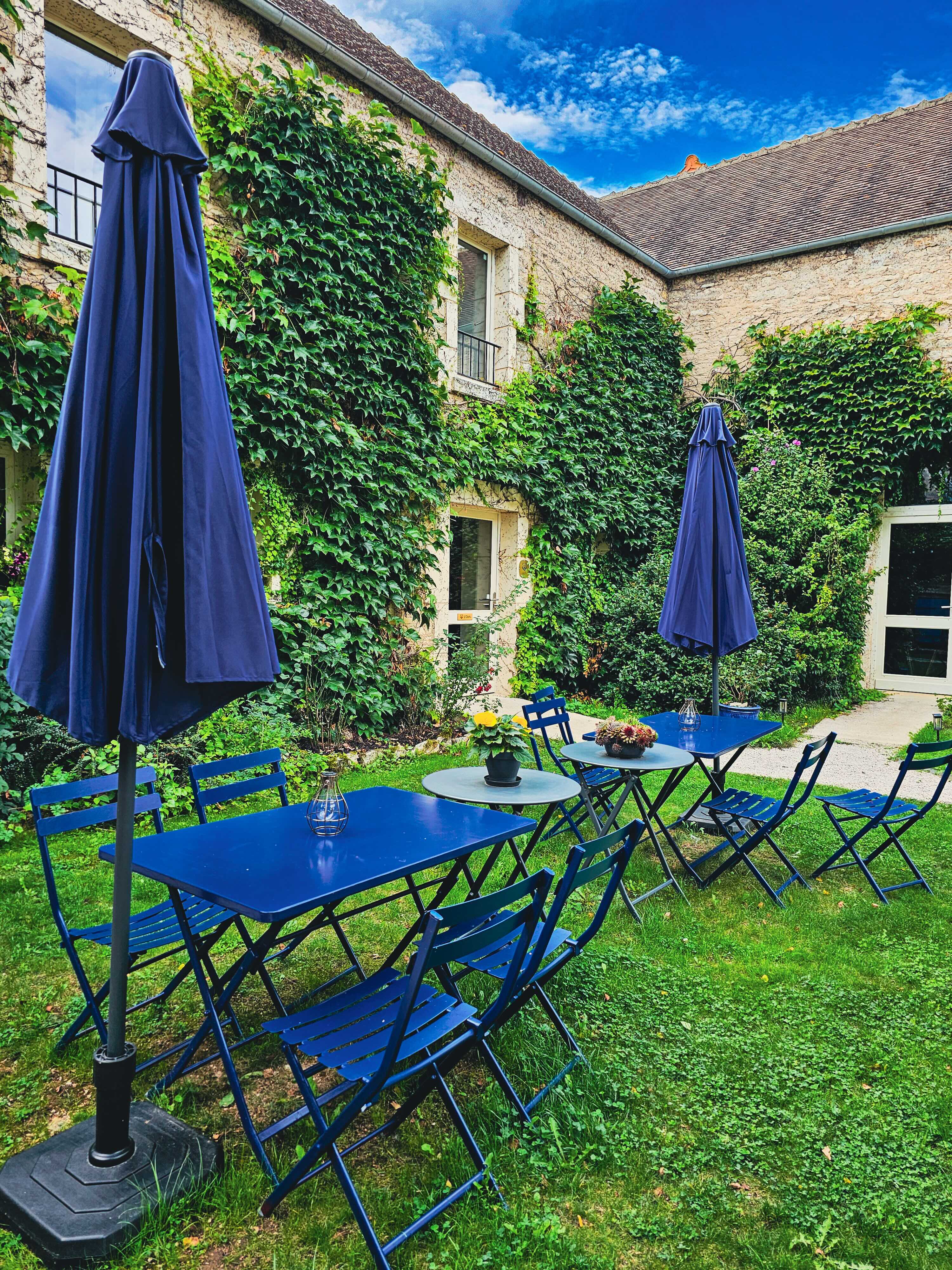 Terrasse jardin avec tables et chaises bleues, parasols et façade couverte de lierre