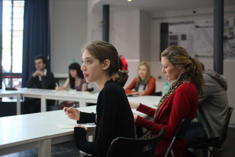 A photo of a group of students listening to a lecture and taking notes while sitting at a table.