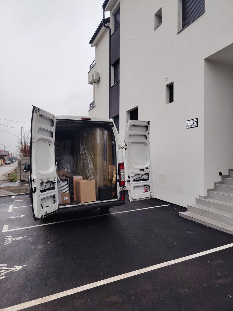 Kombi za selidbe parkiran ispred stambene zgrade, sa otvorenim vratima i pažljivo upakovanim nameštajem i kutijama spremnim za istovar.