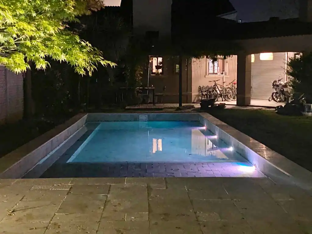 Pileta de hormigón iluminada durante la noche en el patio trasero de una casa, destacando el reflejo del agua celeste en contraste con el entorno.
