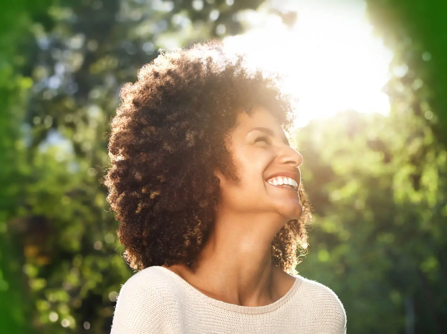 Smiling woman with curly hair in sunlight surrounded by nature.