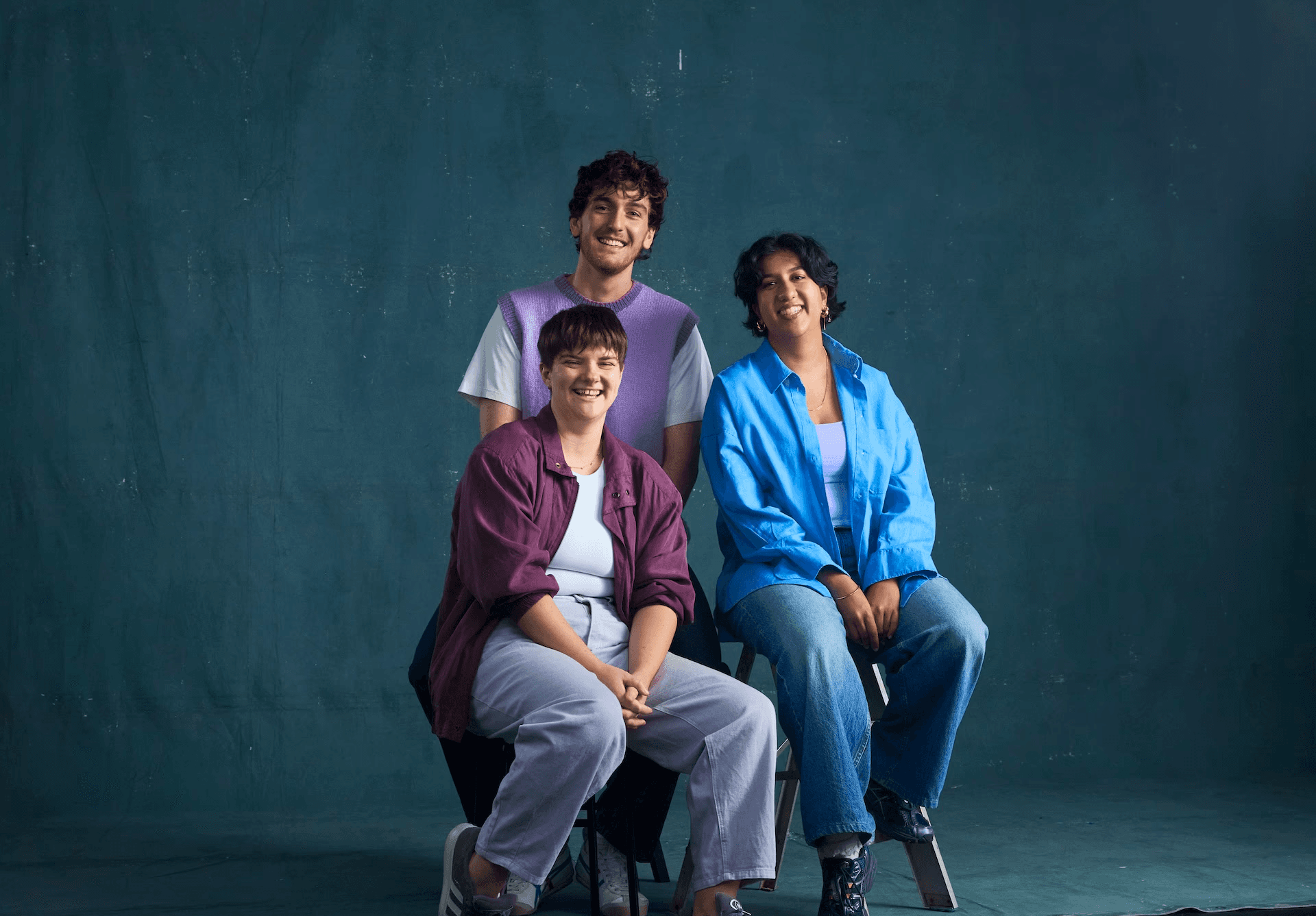 Three young adults in casual, colourful clothing sit and stand against a textured blue studio backdrop. Two sit on stools at the front, smiling toward the camera, while one stands behind them, also smiling. They appear relaxed and friendly, as though part of a creative ensemble or cast portrait.