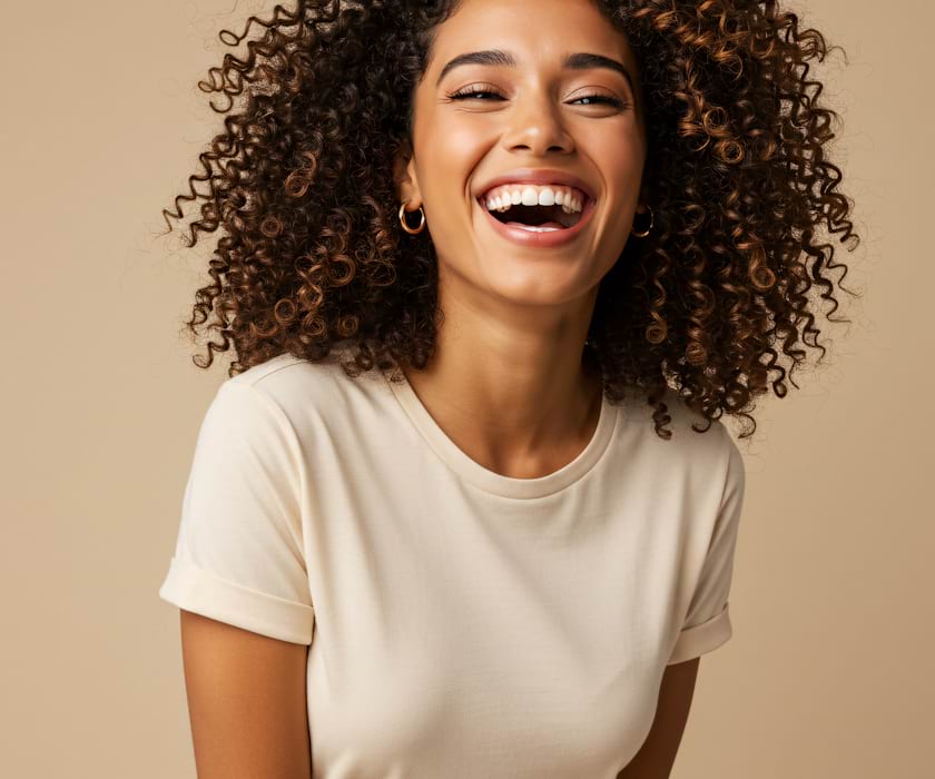 Smiling woman with curly hair wearing a beige t-shirt and pants. The background is a matching beige, creating a warm and joyful atmosphere.