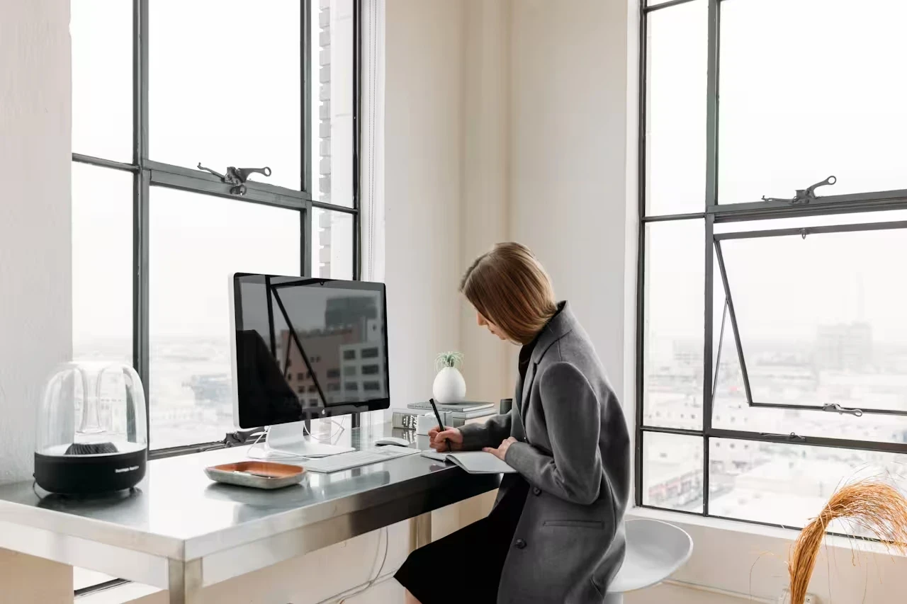 A woman sits alone at a desk by large windows, writing in a notebook beside a desktop monitor.