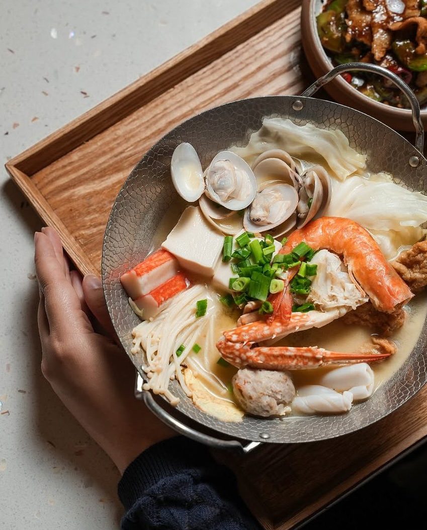 A hand holding a bowl of seafood hotpot, featuring shrimp, fish balls, crab sticks, clams, tofu, and vegetables, placed on a wooden tray.