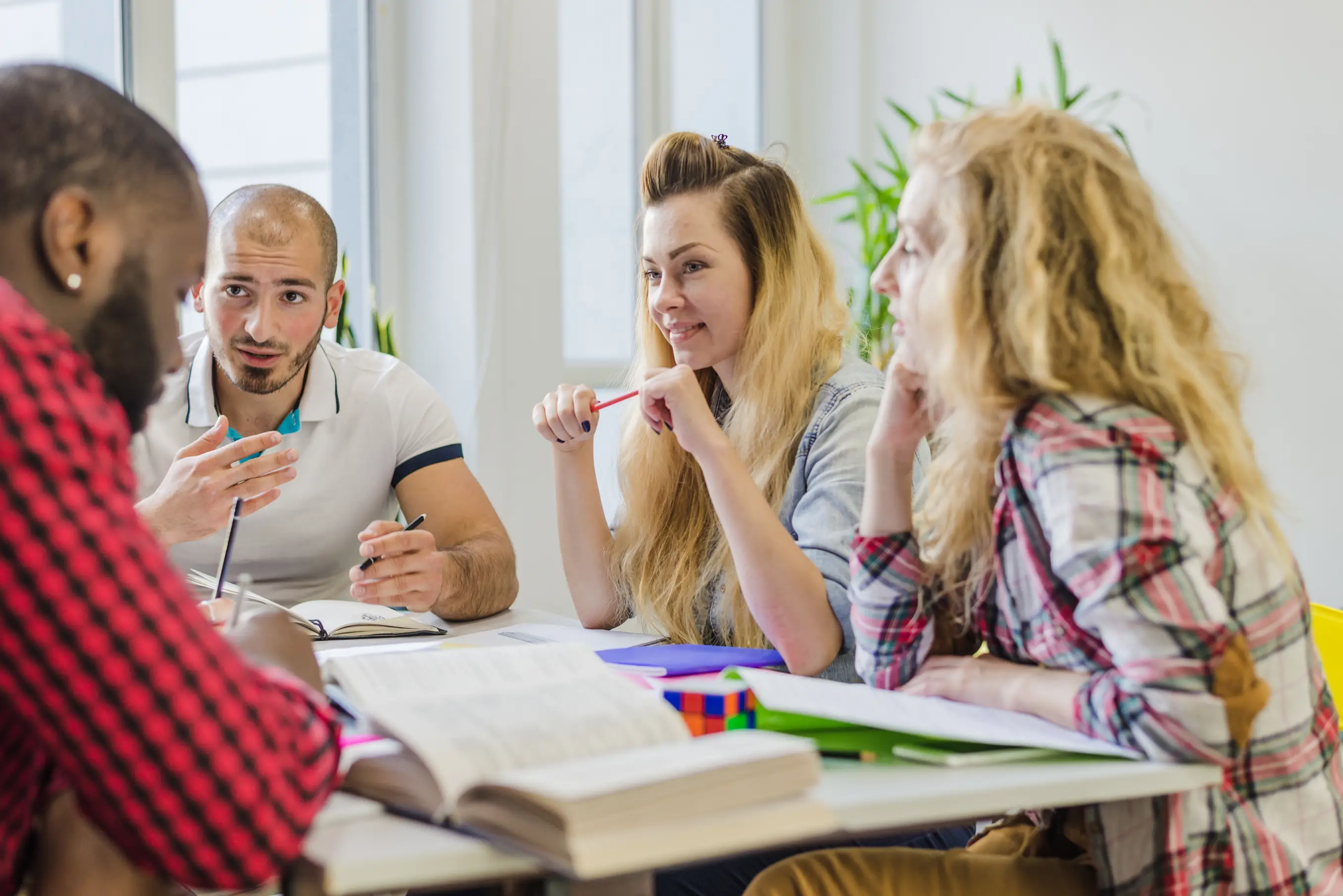 A group of four diverse individuals engaged in a collaborative study session at a table with open books, notebooks, and a colorful multi-layered cube, set in a bright room with plants.