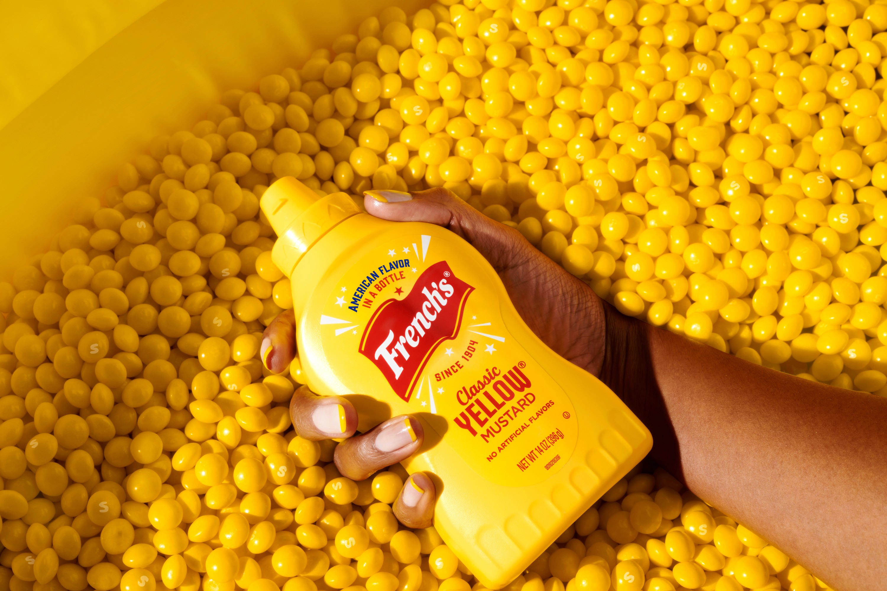 A hand holds a bottle of mustard surrounded by a sea of yellow Skittles candies 