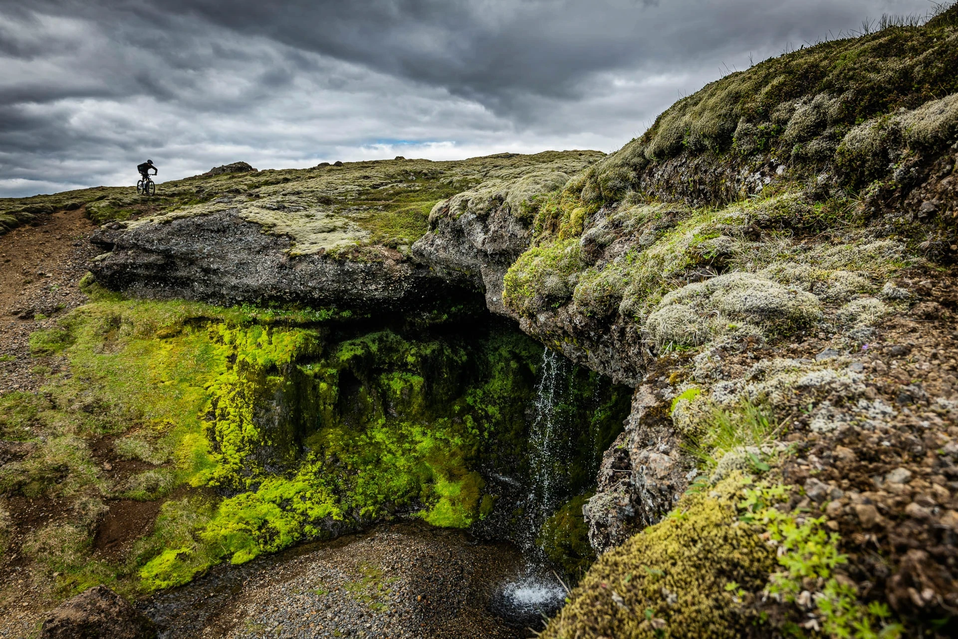 Small waterfall in a mossy canyon, with a cyclist on the trail above.
