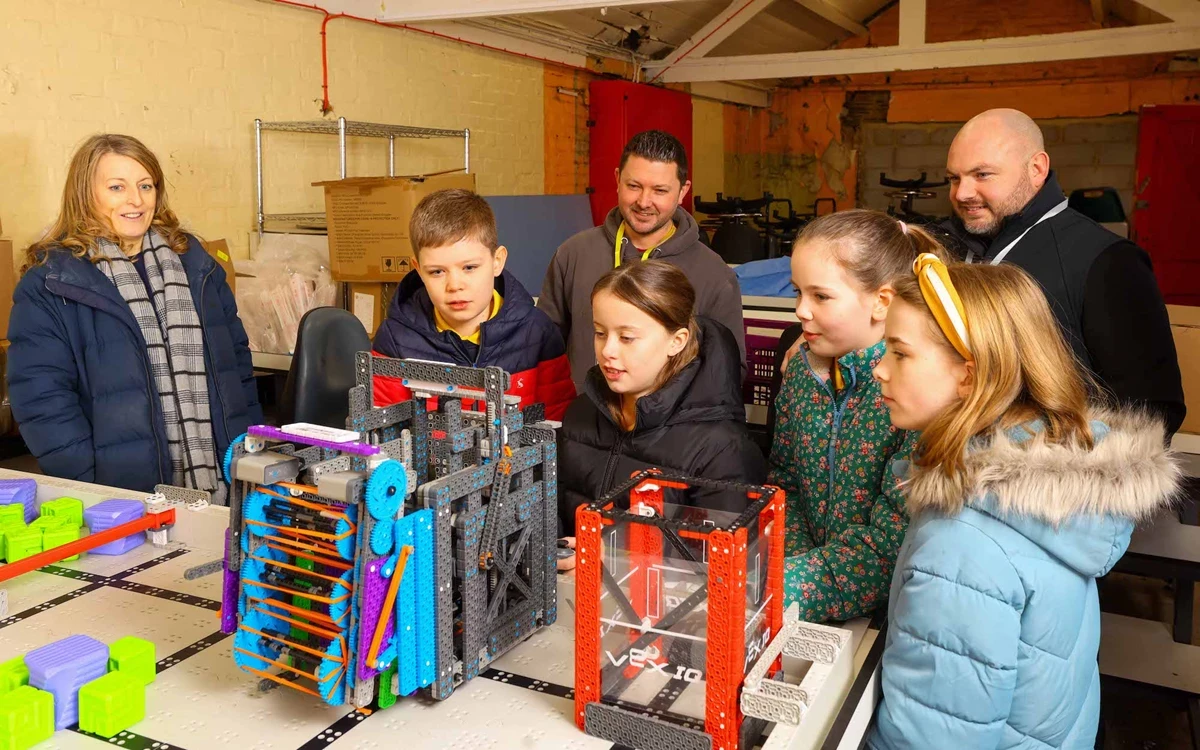 A group of children and adults gather around a robotics competition table inside a workshop-style room with exposed beams and painted brick walls. Several metal-framed robots with colourful components are positioned on the table, along with game pieces arranged in rows. The group appears to be observing or discussing the robots as part of a STEM or robotics activity.