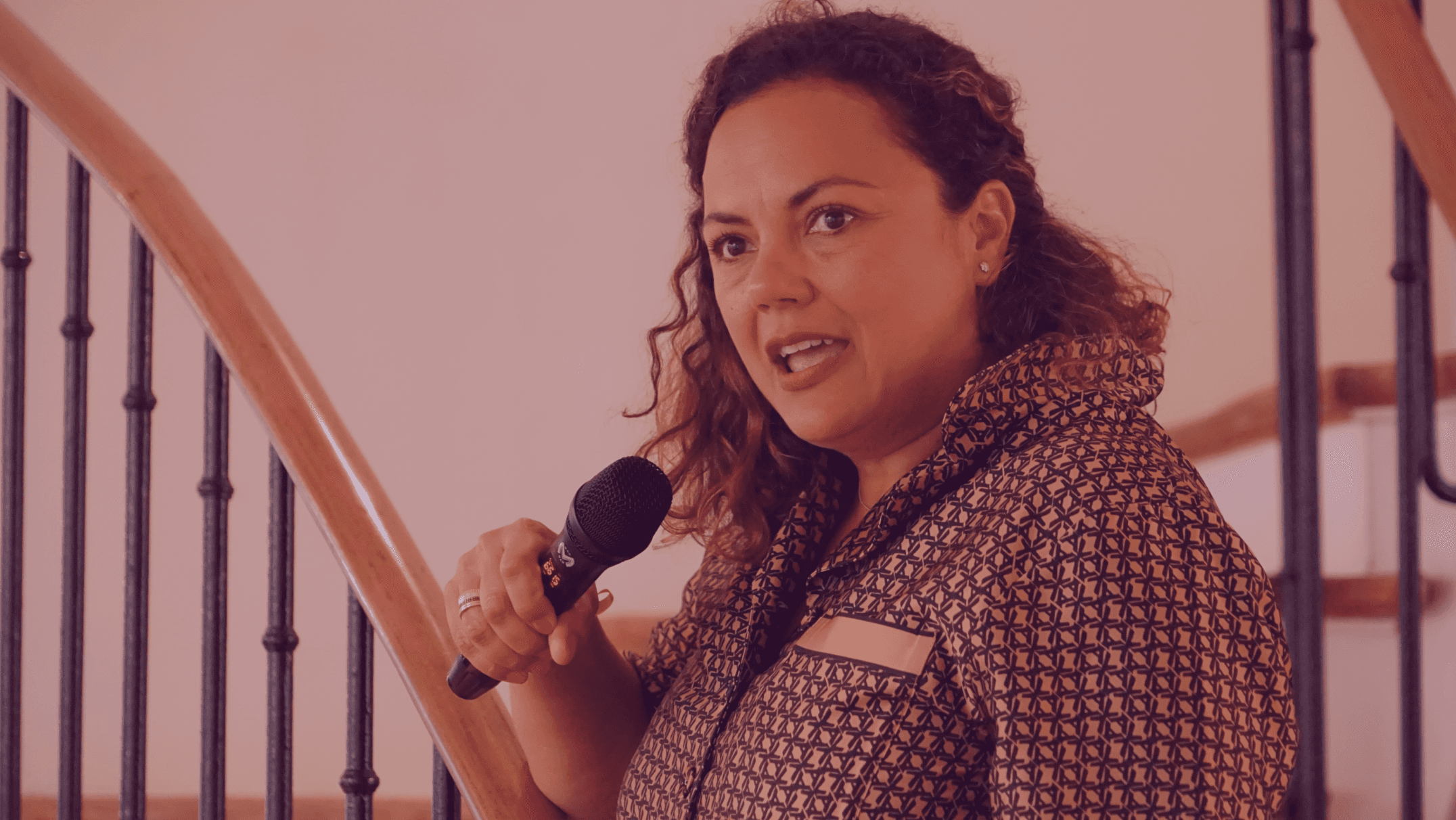 A woman stands near a staircase, holding a microphone and looking towards the camera with a confident expression.
