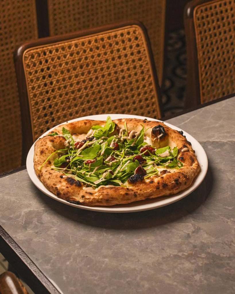 A close-up view of a freshly made pizza topped with arugula, roasted vegetables, and drizzled with sauce, served on a white plate atop a stone table.