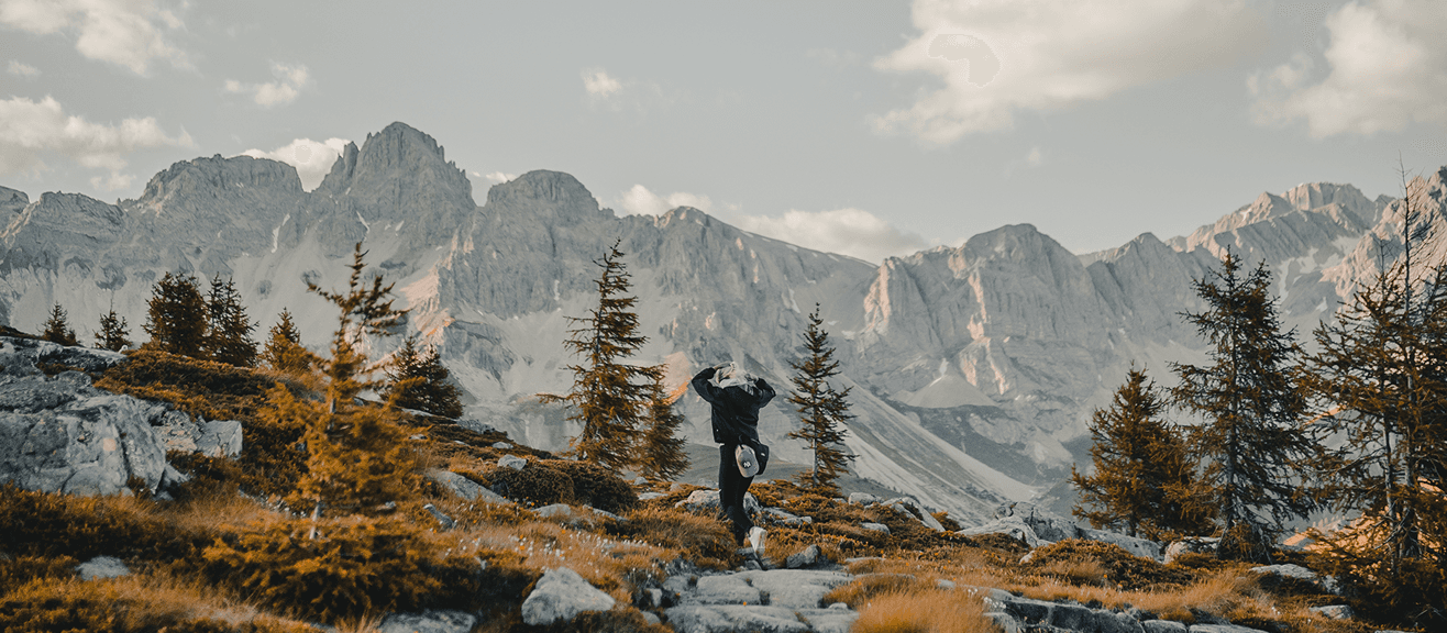 A hiker stands on a rocky mountain trail, looking out at a vast, jagged mountain range in the background.