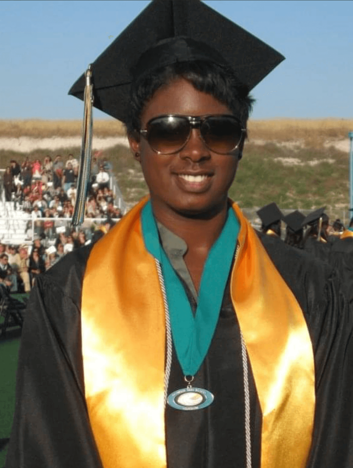 Ashleigh Warren, a graduate wearing a cap and gown, smiling and wearing sunglasses, with a colorful stole around their neck.