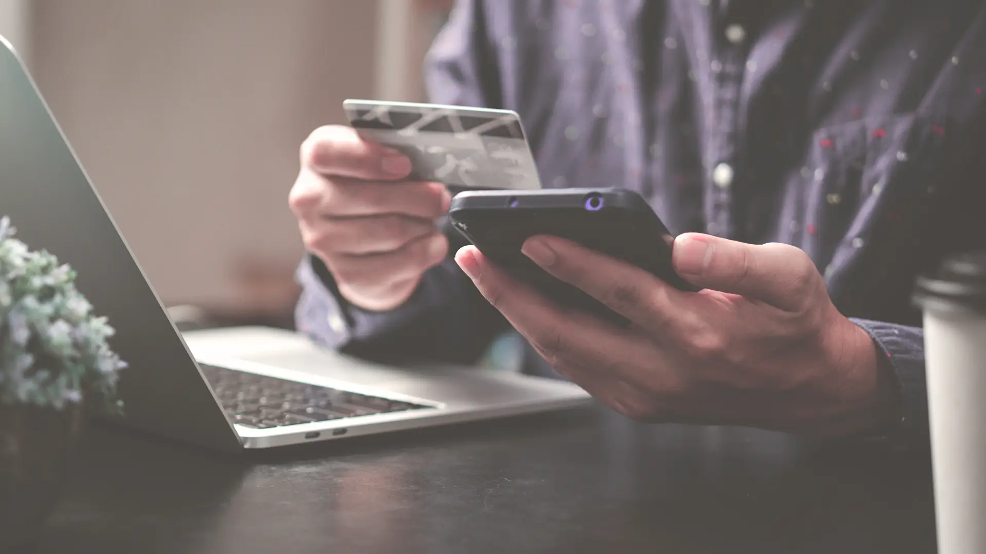 Person holding credit card and smartphone while shopping online with a laptop nearby—illustrating smart and secure online shopping habits in 2025.
