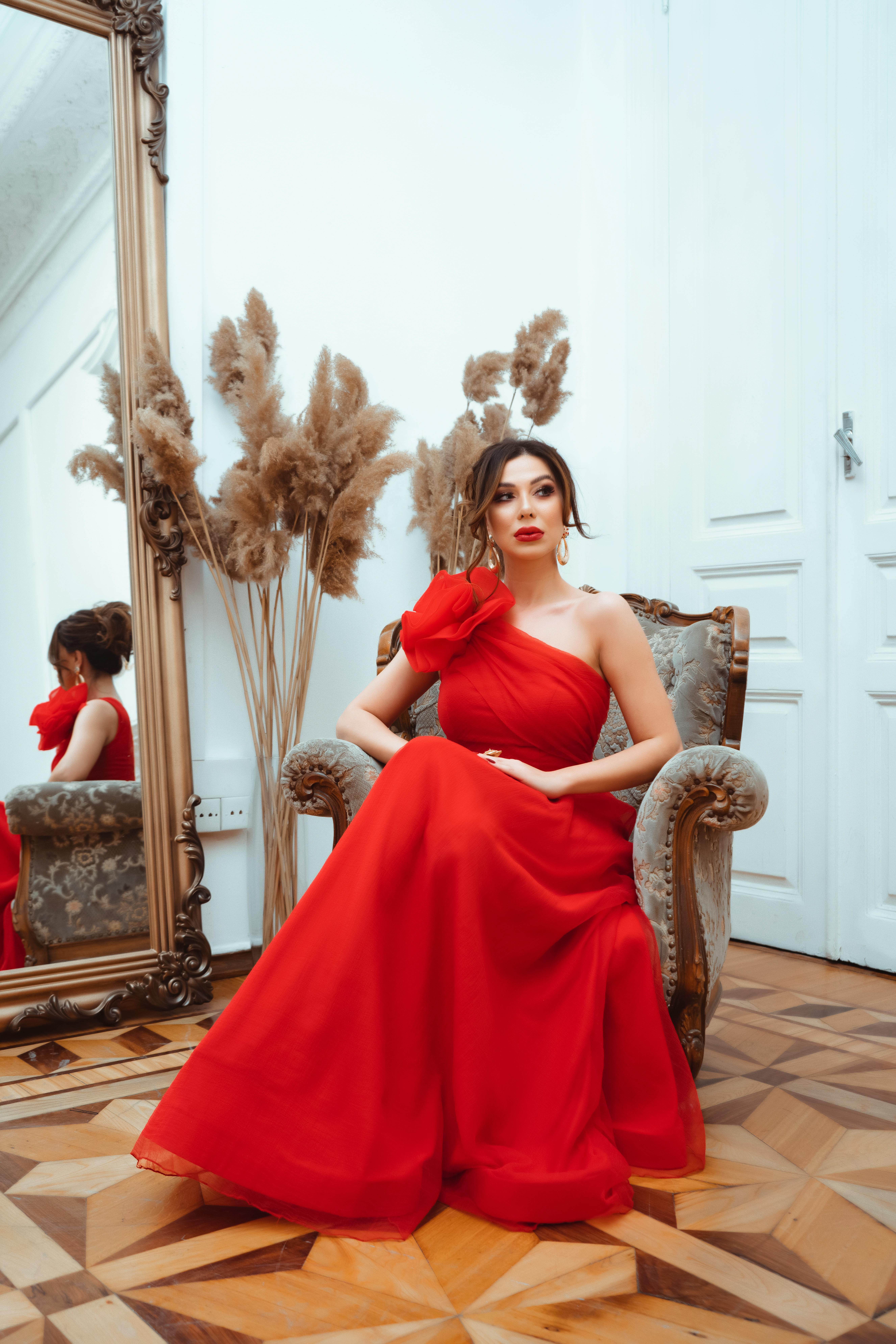 Elegant woman in a red one-shoulder gown sitting on an ornate armchair in a stylish room with a large mirror and dried pampas grass decor.