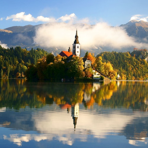 אי עם כנסייה, עצי סתיו והשתקפות על אגם רגוע, על רקע הרים ושמים מעוננים חלקית.