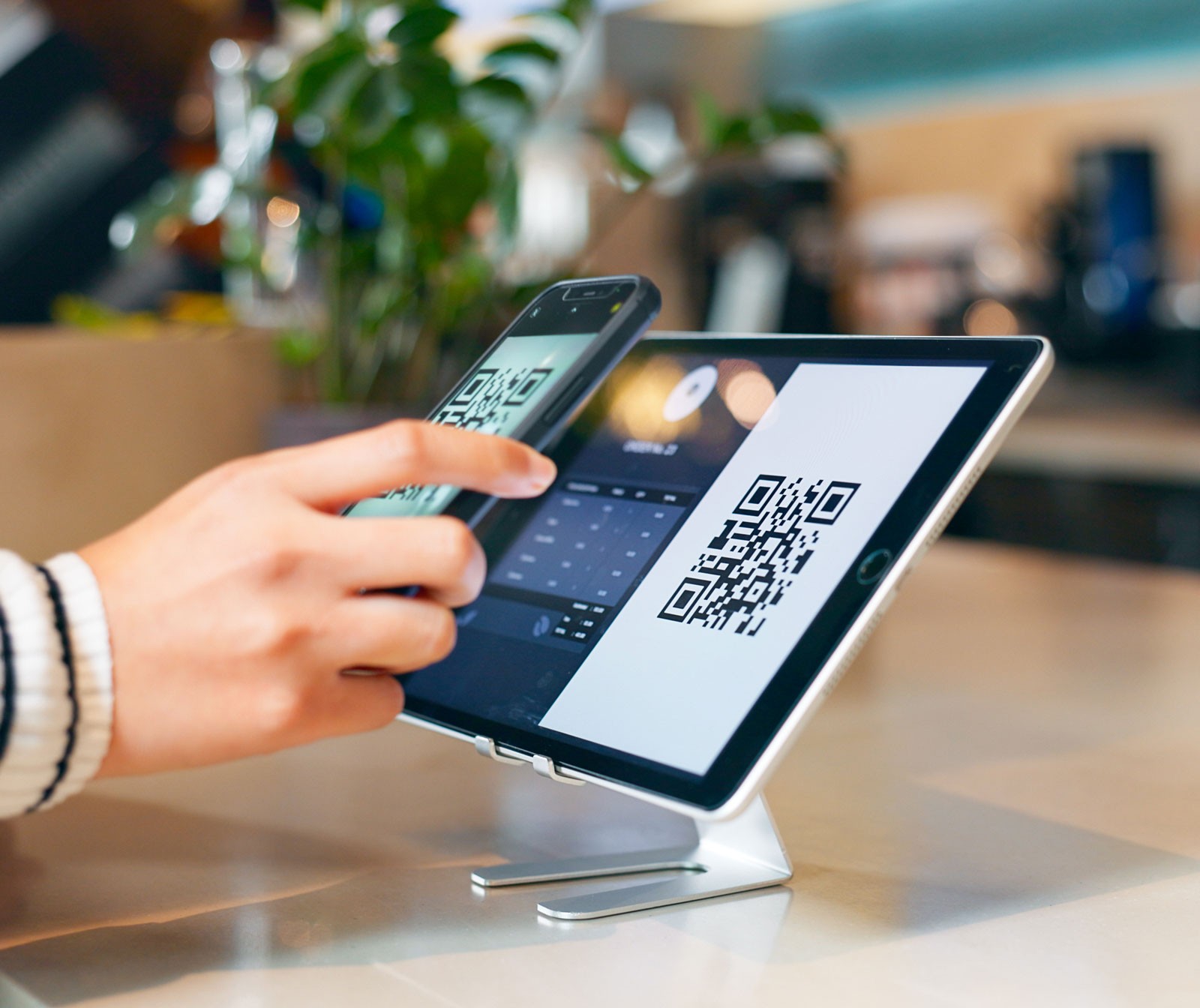 A person using a smartphone to scan a QR code displayed on a tablet screen at a checkout counter, representing digital payment technology and contactless transactions.