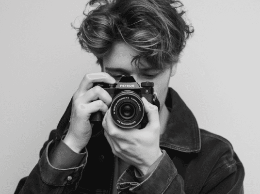 A person with tousled hair is holding a vintage camera up to their face, wearing a dark denim jacket in a black and white photograph.