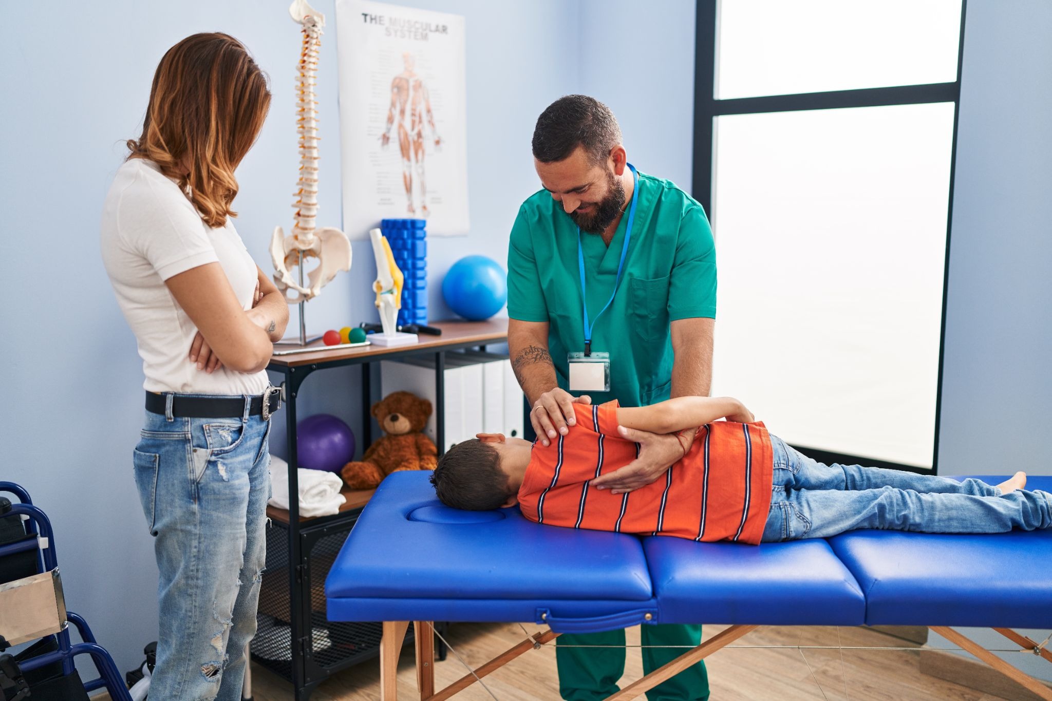 A male physiotherapist examining a young boy lying on a treatment table while his mother watches.