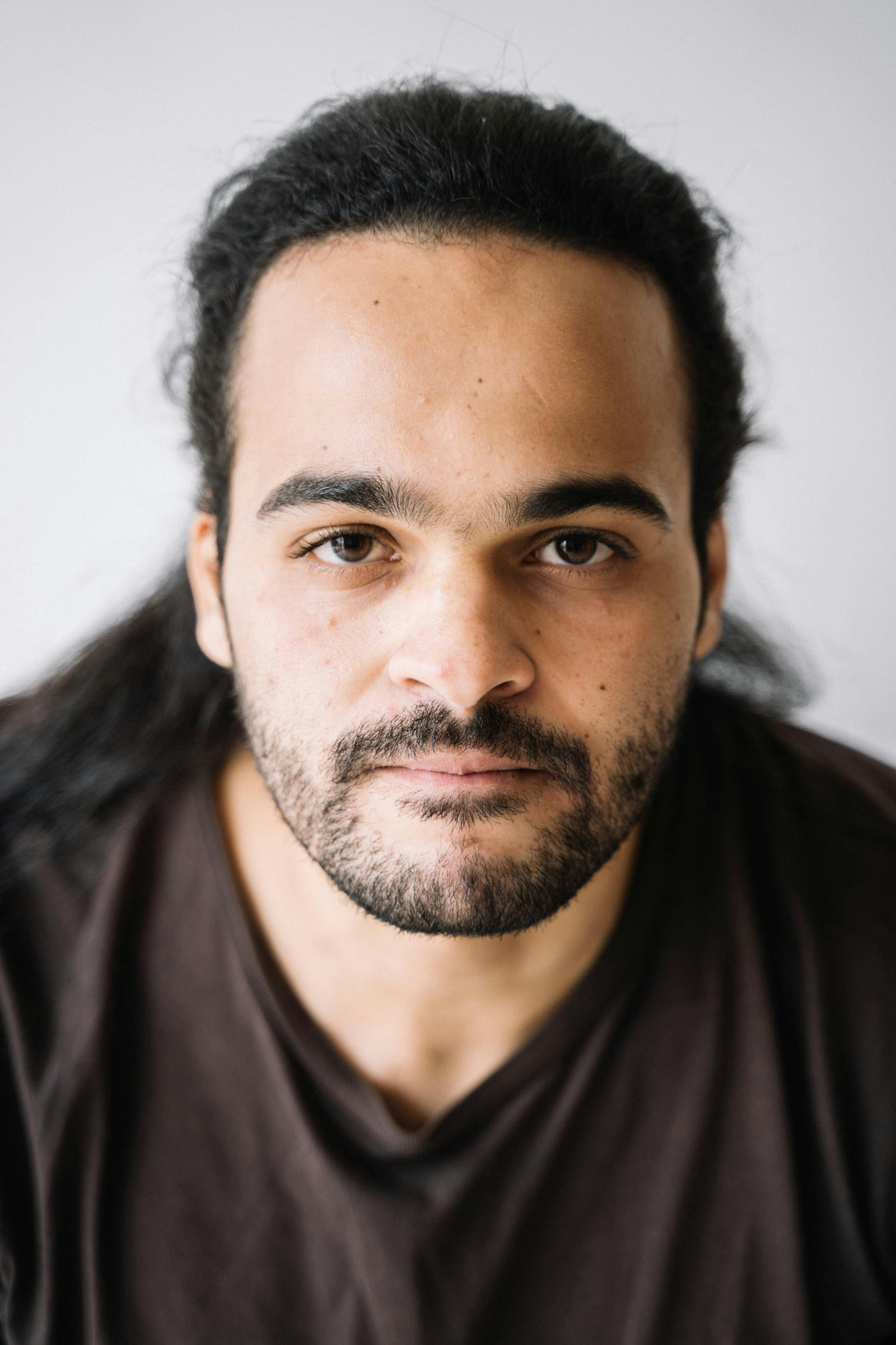 A man with long dark hair and a beard gazes directly at the camera with a neutral expression. He wears a brown shirt against a plain white background.
