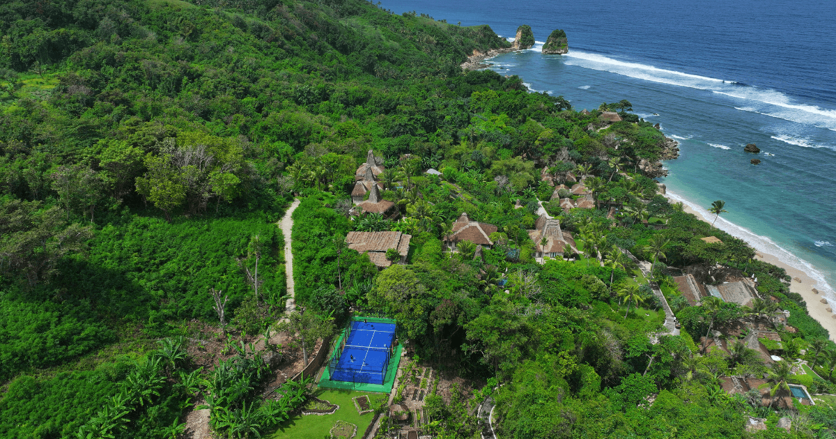 A view of a resort and a blue padel court near the ocean surrounded by lush greenery