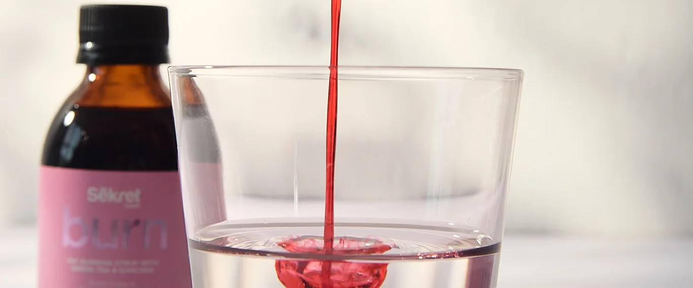 Red Sēkret Burn syrup poured into a glass of water beside its bottle