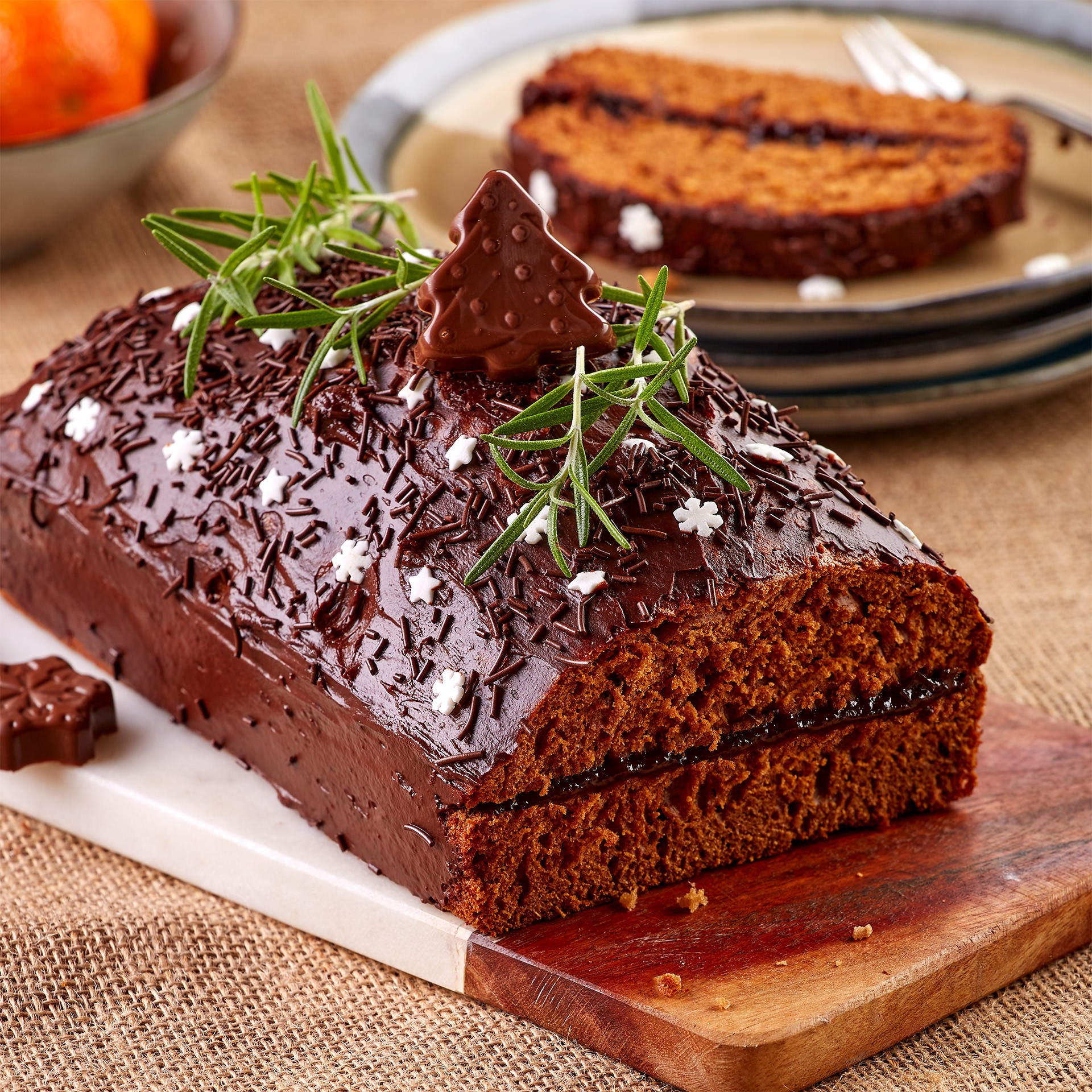A rich, chocolate-glazed gingerbread loaf topped with sprigs of rosemary and adorned with snowflake and tree-shaped decorations is presented on a wooden serving board, with a plate of sliced gingerbread in the background.