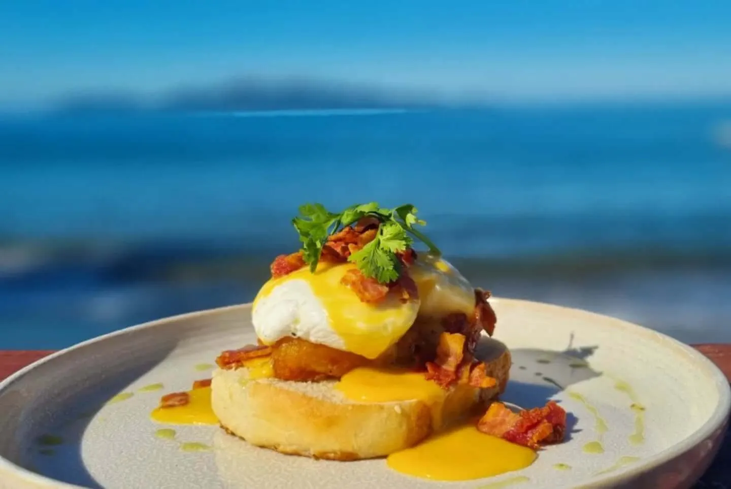 Delicious Eggs Benedict with bacon, hollandaise, and herbs, served on a plate against a vibrant Fiji beachfront.