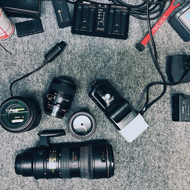 A flat lay of camera equipment including lenses, a flash, chargers, batteries, memory cards, and cables arranged on a gray surface.